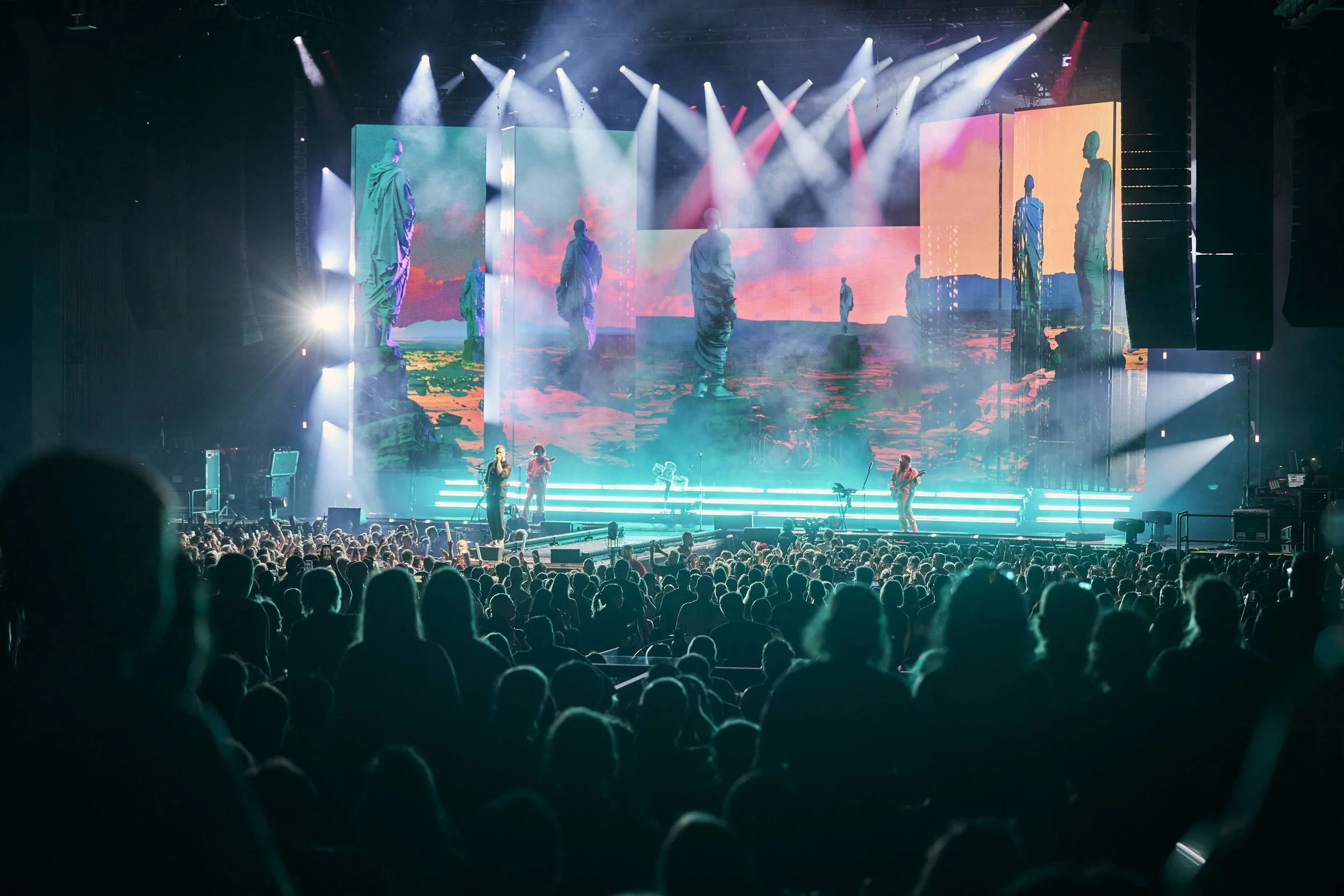 A live concert in an indoor arena with a large audience and a brightly lit stage featuring multiple screens displaying scenic landscape images and silhouettes of performers.