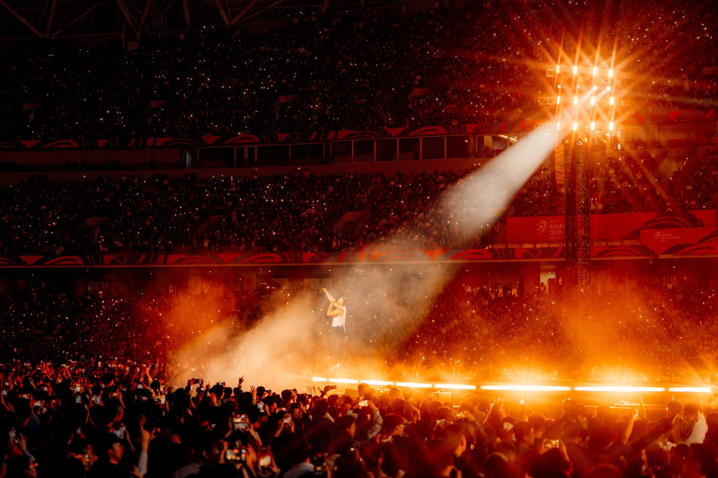 A performer singing on stage with a microphone under a bright spotlight, surrounding smoke effects, in front of a large audience in a stadium filled with seated spectators, at night.