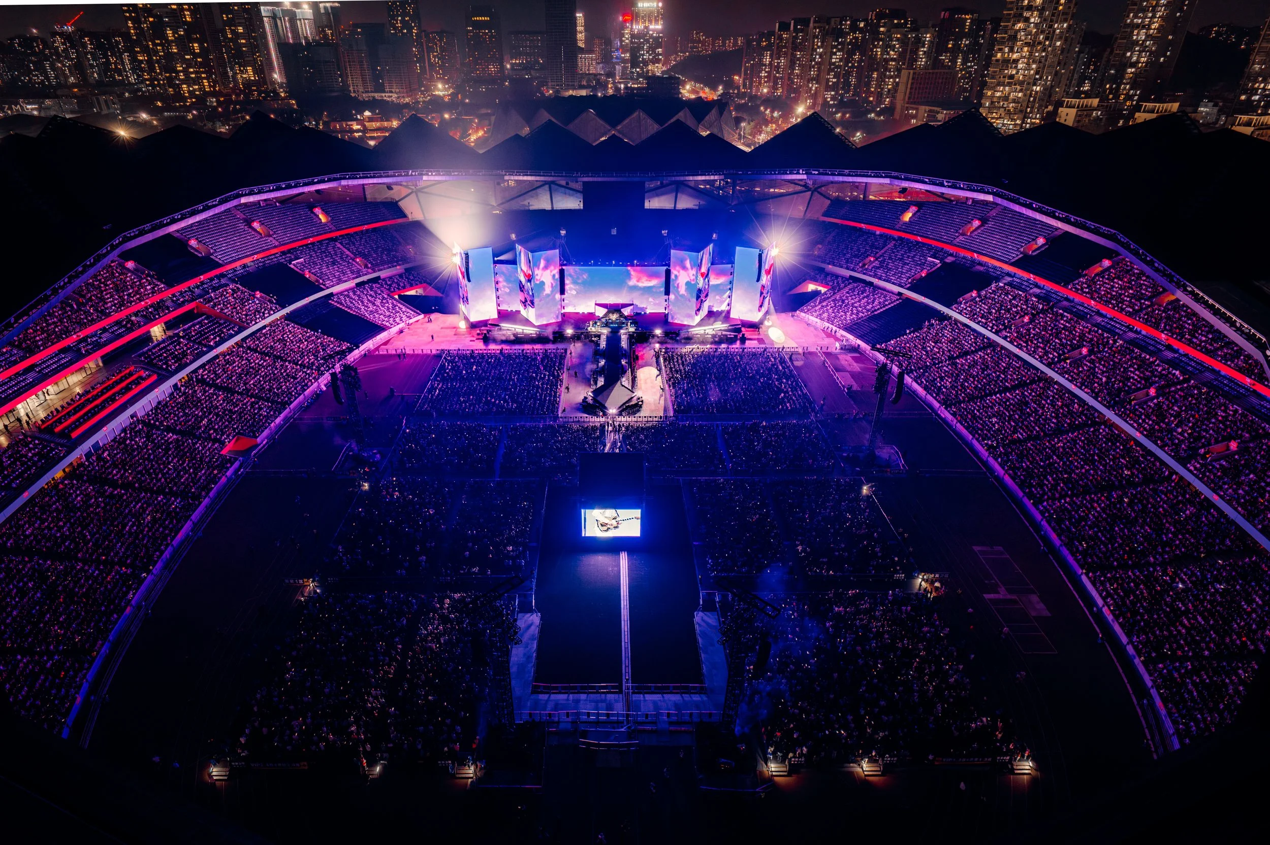 Aerial view of a large outdoor concert stadium at night, illuminated with purple and blue lights, filled with a crowd of people, with stage visuals and city skyline in the background.