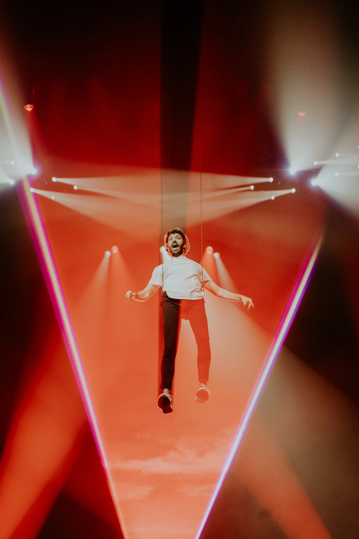 Performer suspended in the air on stage with dramatic red laser lighting and beams of light overhead.