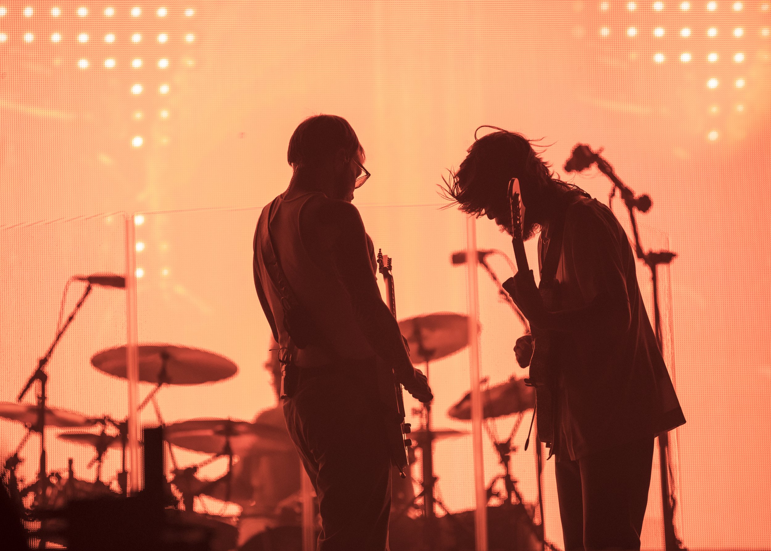 Silhouettes of two musicians playing electric guitars on stage with a vibrant orange background and drums in the background.