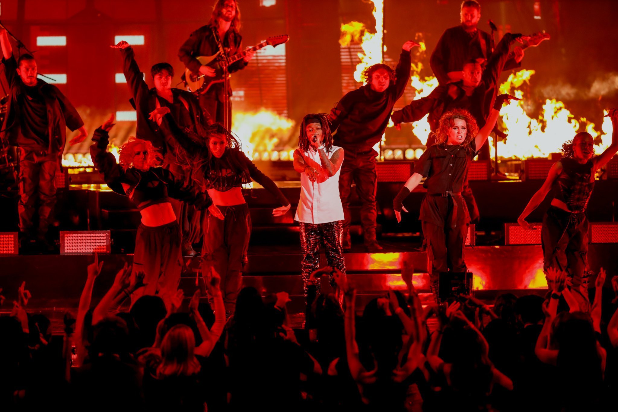 Performer singing on stage surrounded by dancers with flames in the background and an audience in front.
