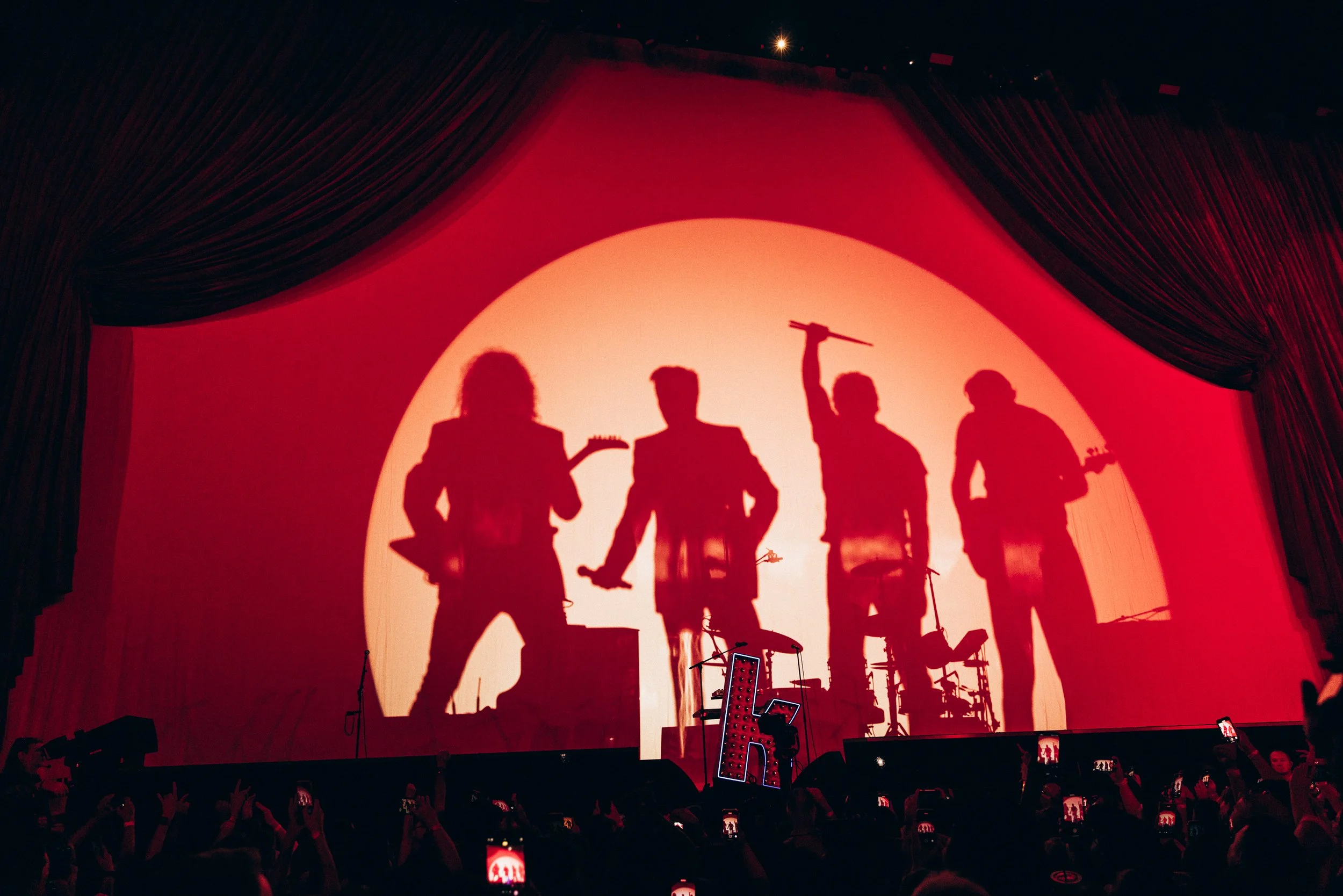 Silhouettes of four band members performing on stage with a large red circular backdrop behind them.