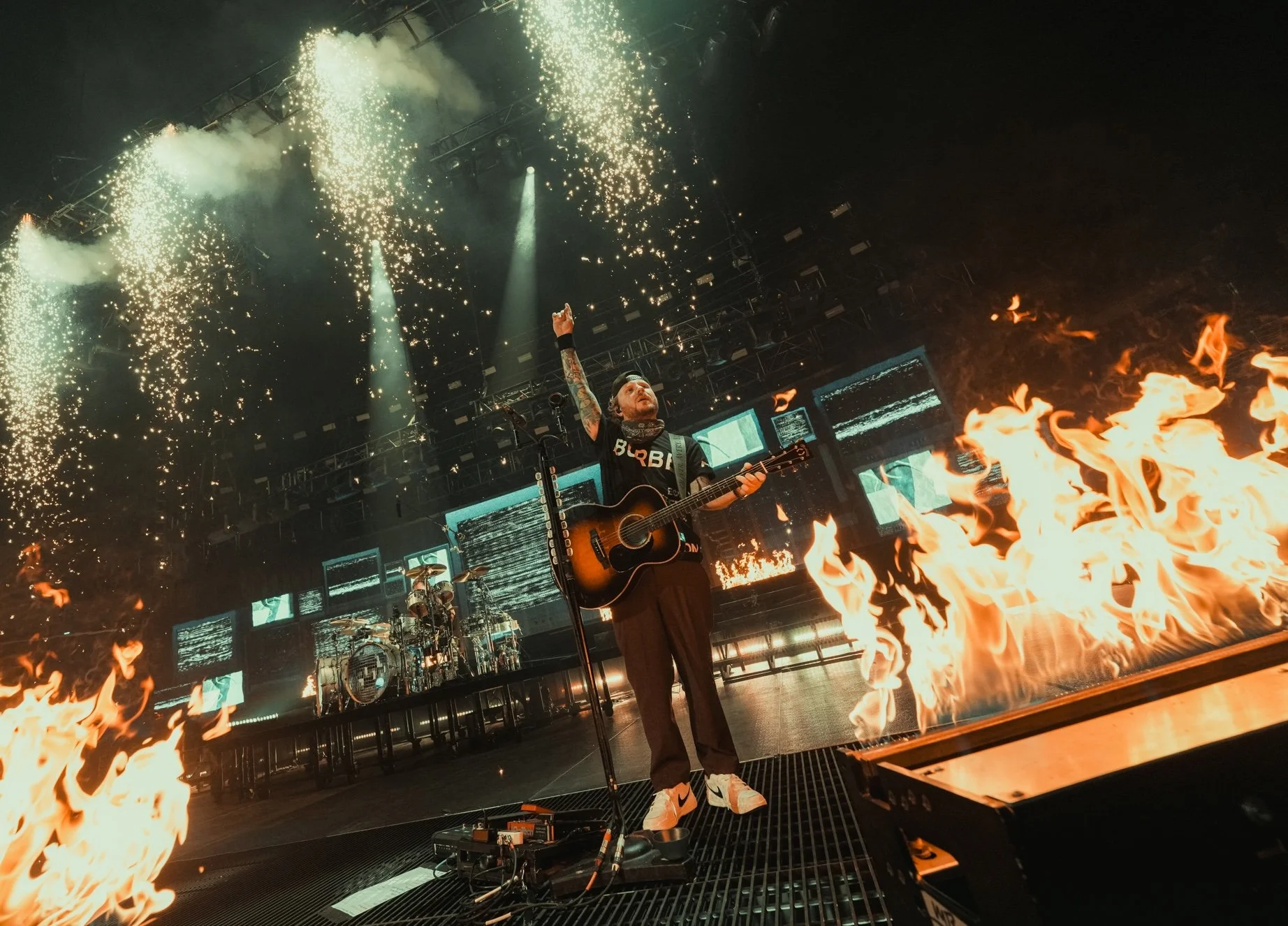A musician playing an acoustic guitar during a concert with fire and pyrotechnics, with sparks and flames on stage.