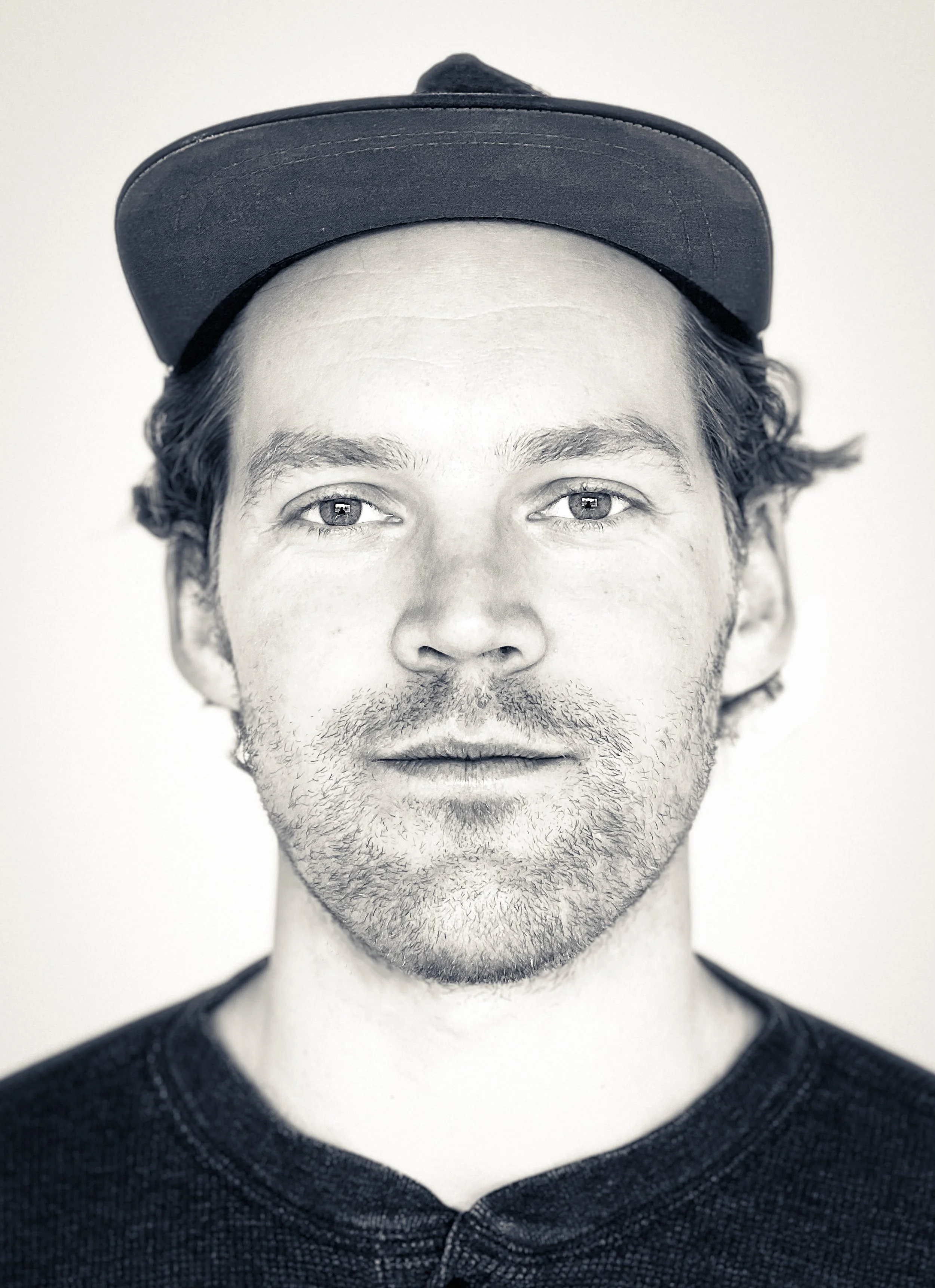 Black and white portrait of a man wearing a baseball cap, with short, curly hair, light facial hair, and a neutral expression.