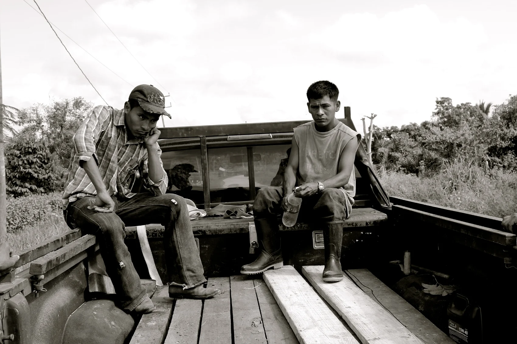 Two young men sitting on the back of a pickup truck in a rural area. One is wearing a plaid shirt and a cap, resting his chin on his hand. The other wears a sleeveless shirt and boots, holding a water bottle. There are trees and power lines in the ba