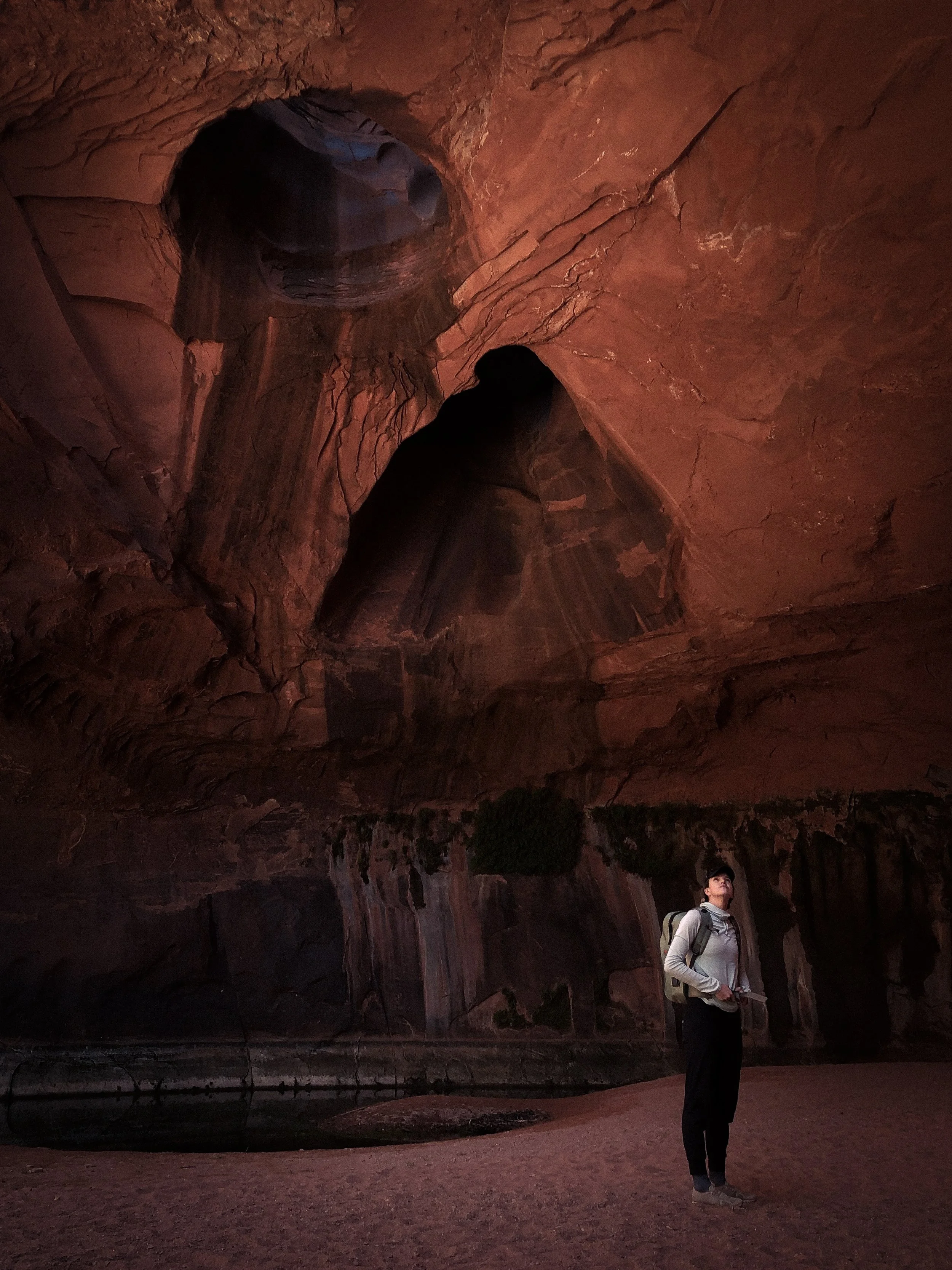 A person standing inside a large canyon with red rock walls, a small pool of water at the base, and interesting rock formations above.