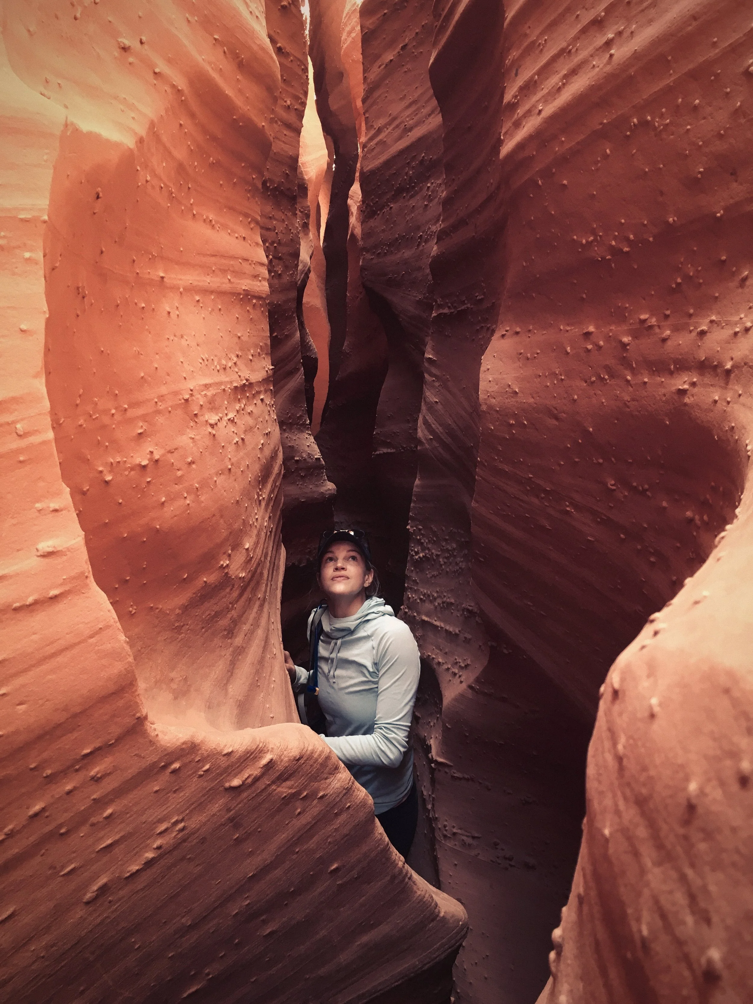 A woman standing in a narrow slot canyon with smooth, layered red rock walls surrounding her.