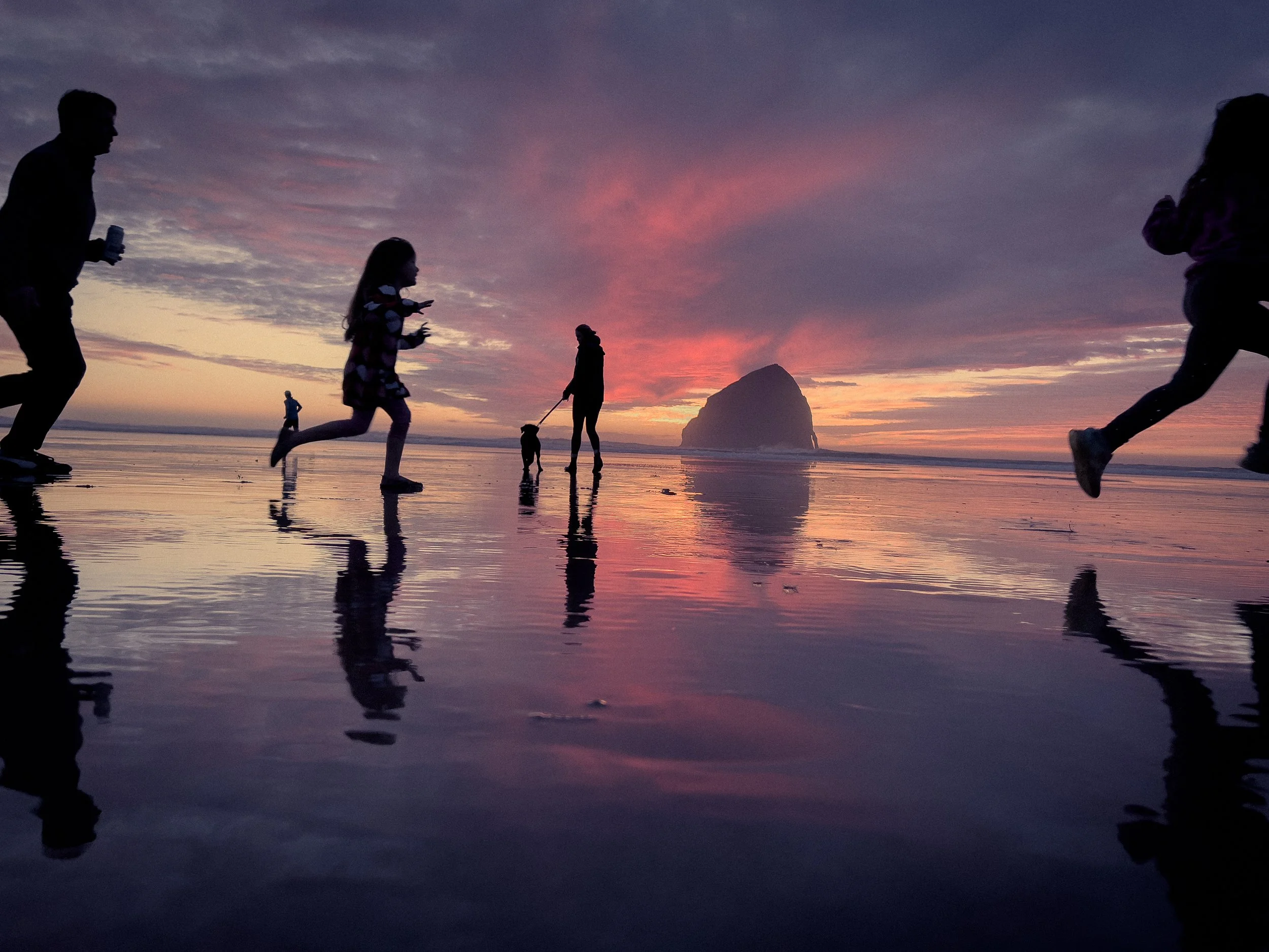 Silhouettes of children and a woman playing on wet sand during a colorful sunset at the beach, with an island in the background.