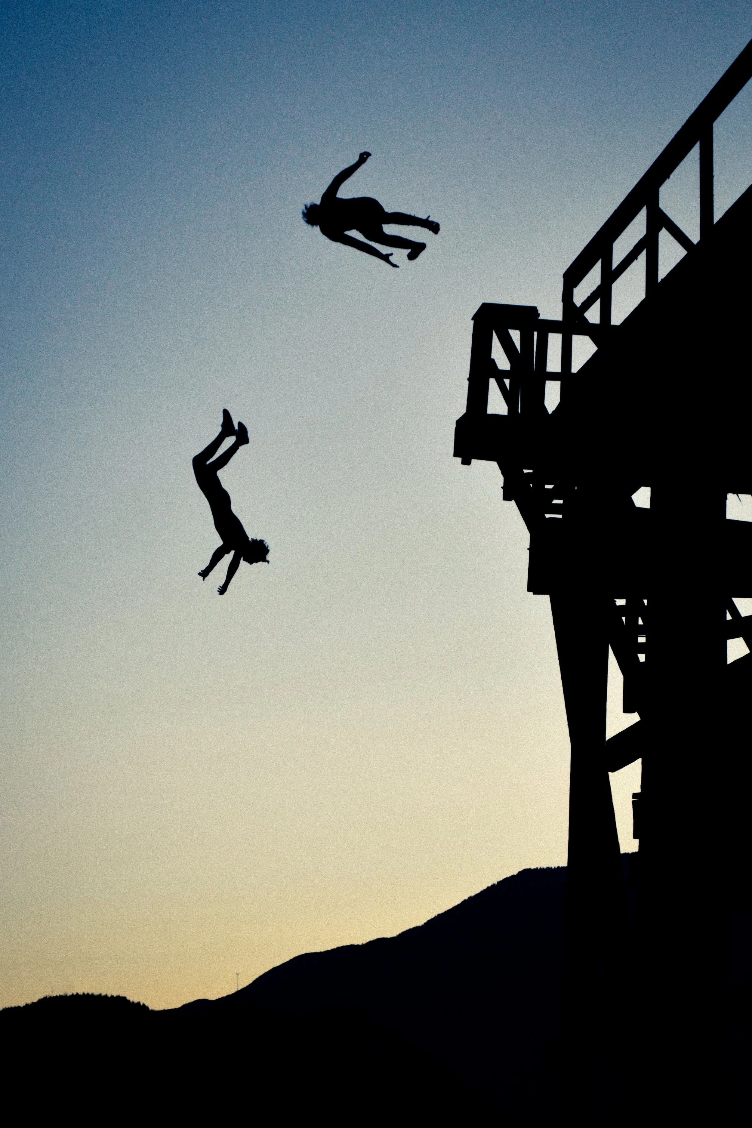 Two silhouetted people in mid-air jump off a pier against a dusky sky, with mountains in the background.