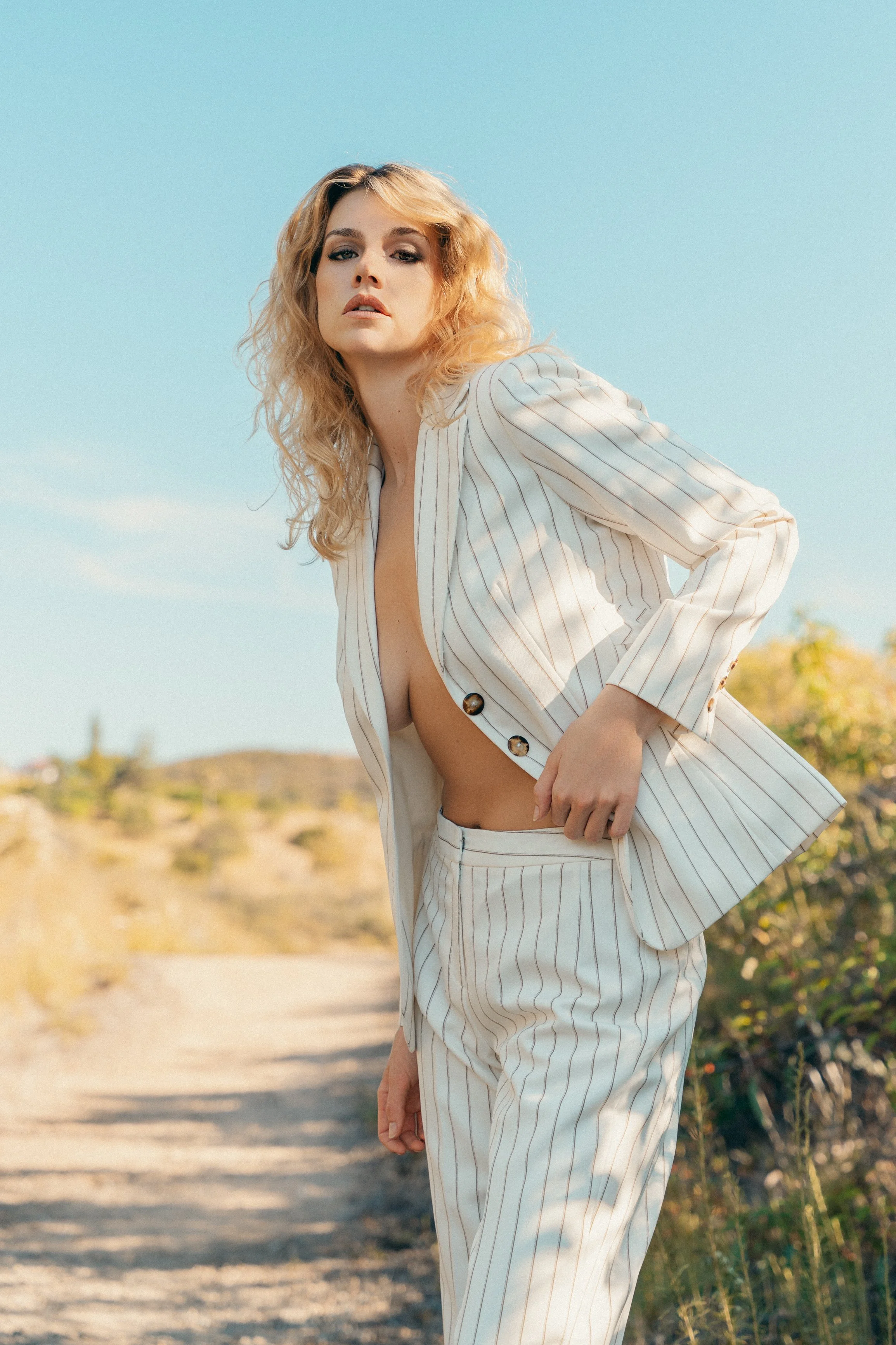 A woman in a striped suit with the jacket unbuttoned, revealing her midriff, standing on a dirt path outdoors with a clear blue sky in the background.