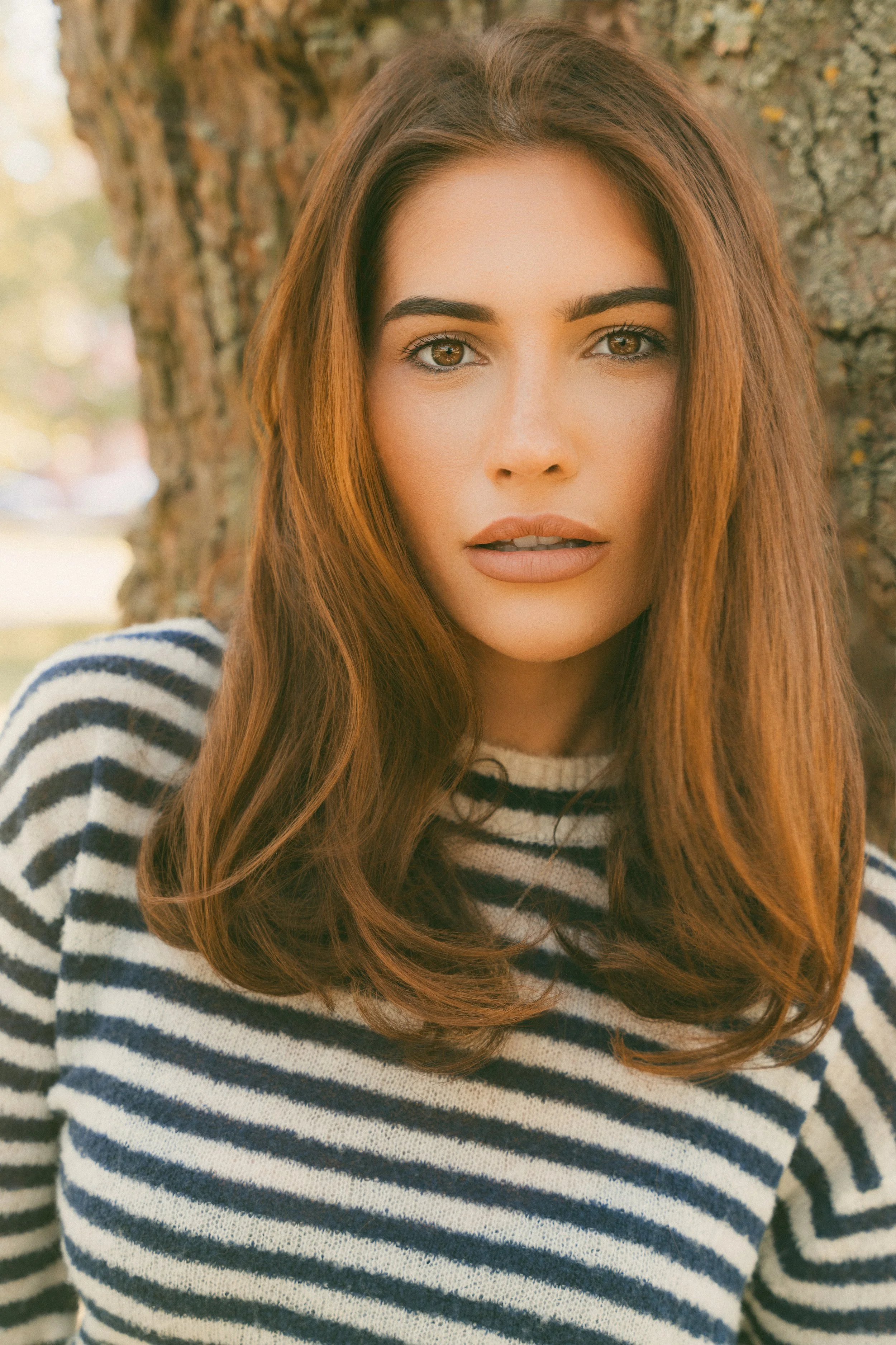 A young woman with long, wavy auburn hair and green eyes standing outdoors near a tree, wearing a striped black and white sweater.