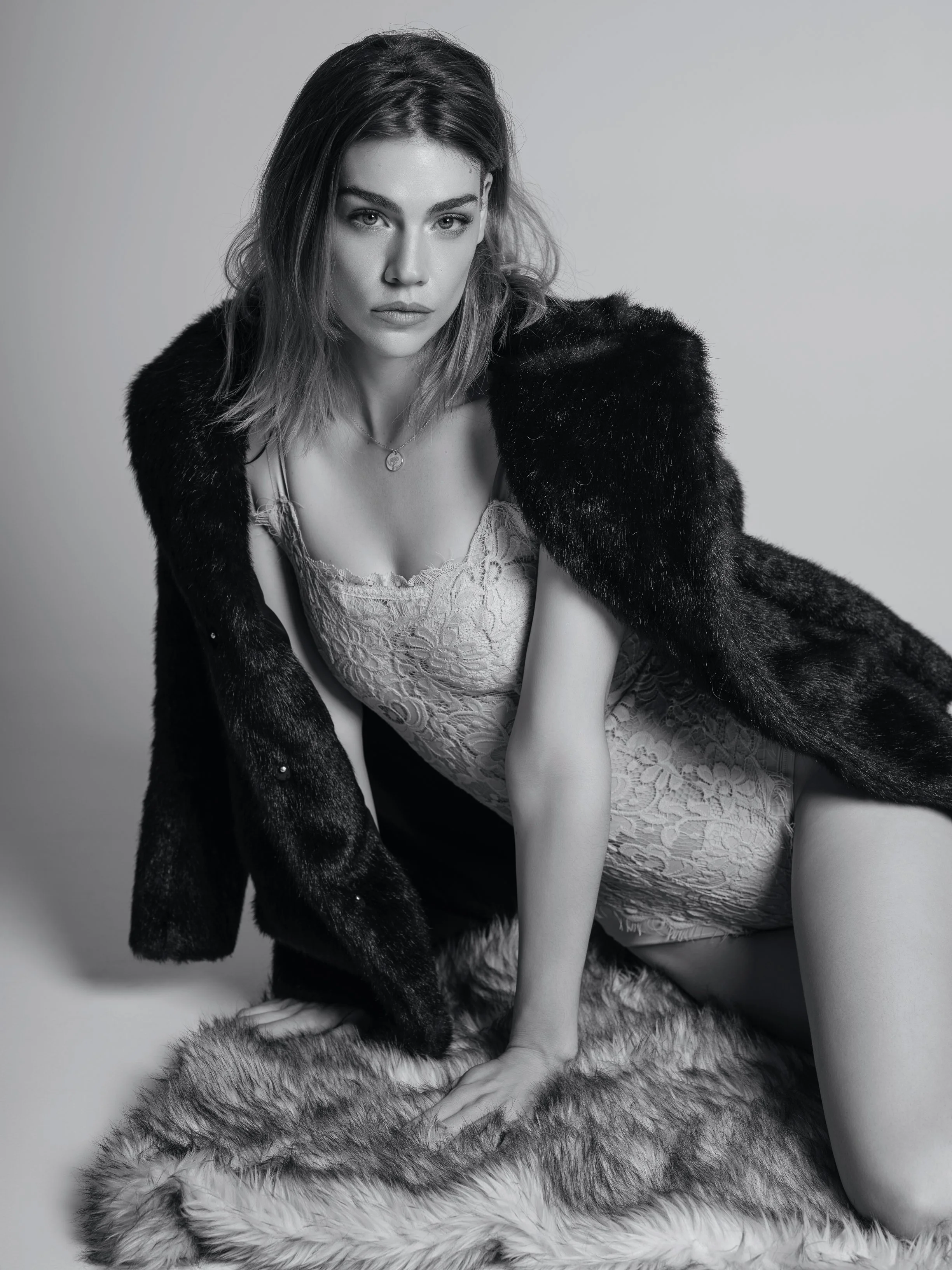 A woman with medium-length hair wearing a lace dress, a necklace, and a fur coat, kneeling on a furry rug, in black and white studio portrait.