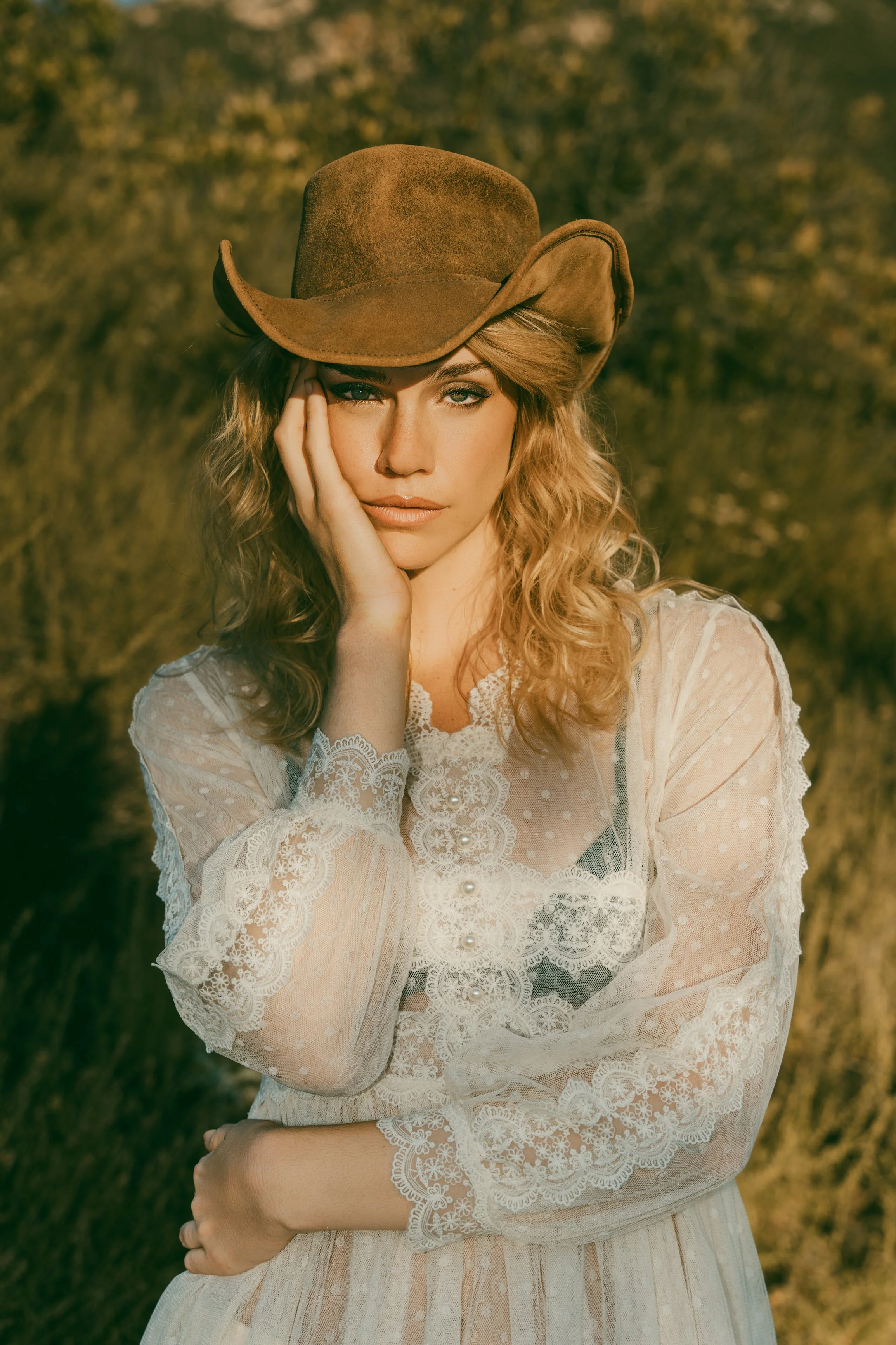 A woman with wavy blonde hair wearing a brown cowboy hat and a white lace dress, standing outdoors with a blurred natural background.