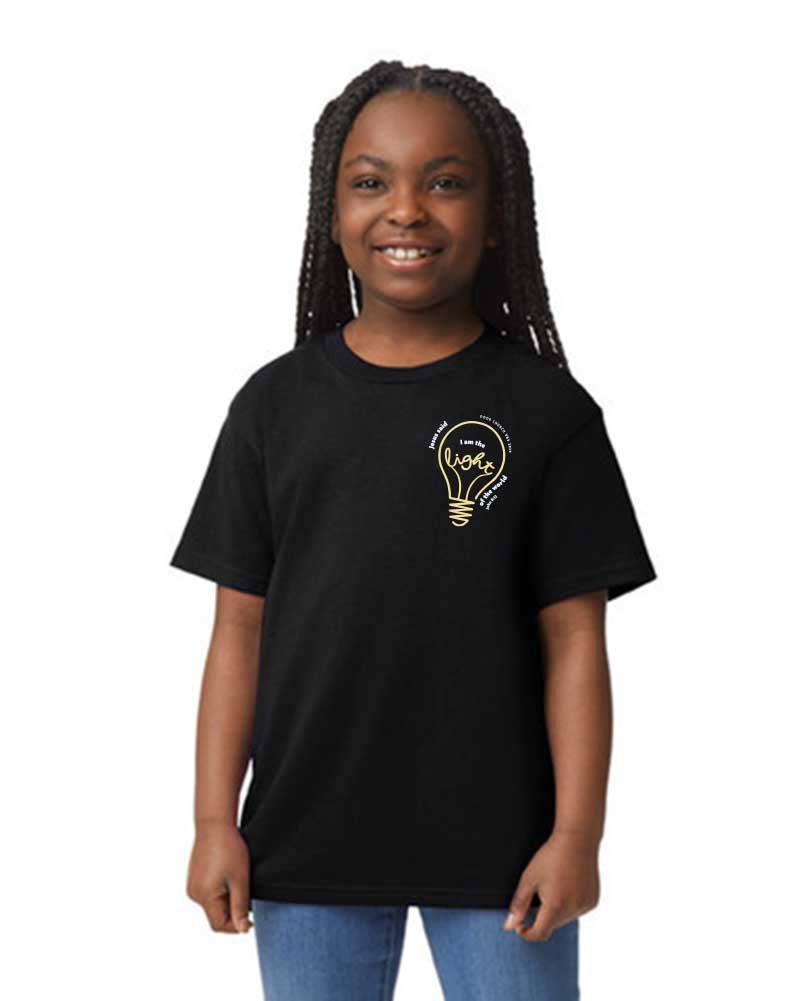 A young girl with braided hair smiling, wearing a black t-shirt with a yellow lightbulb graphic and text, standing against a plain white background.
