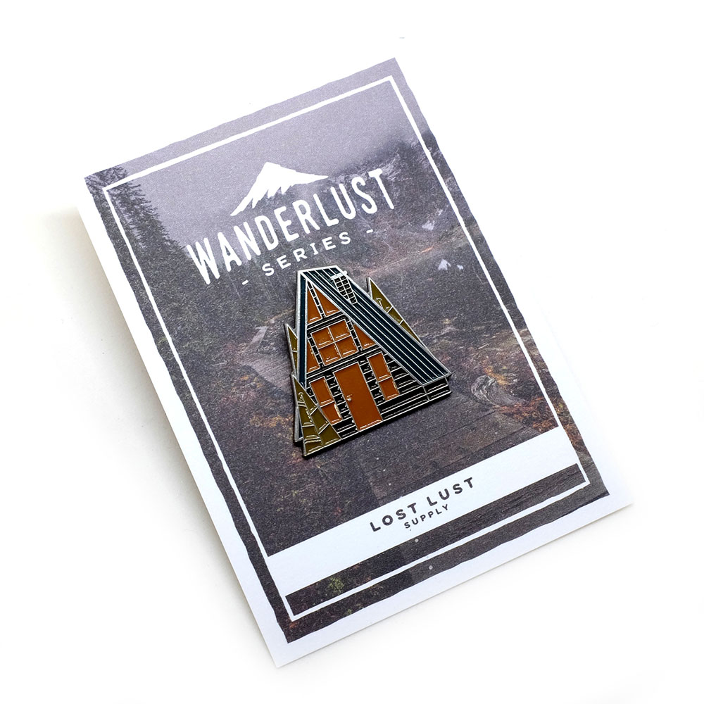 Rear view of die-cast soft enamel pin of an A-frame cabin in the Spring and full color backing card, by Lost Lust Supply.