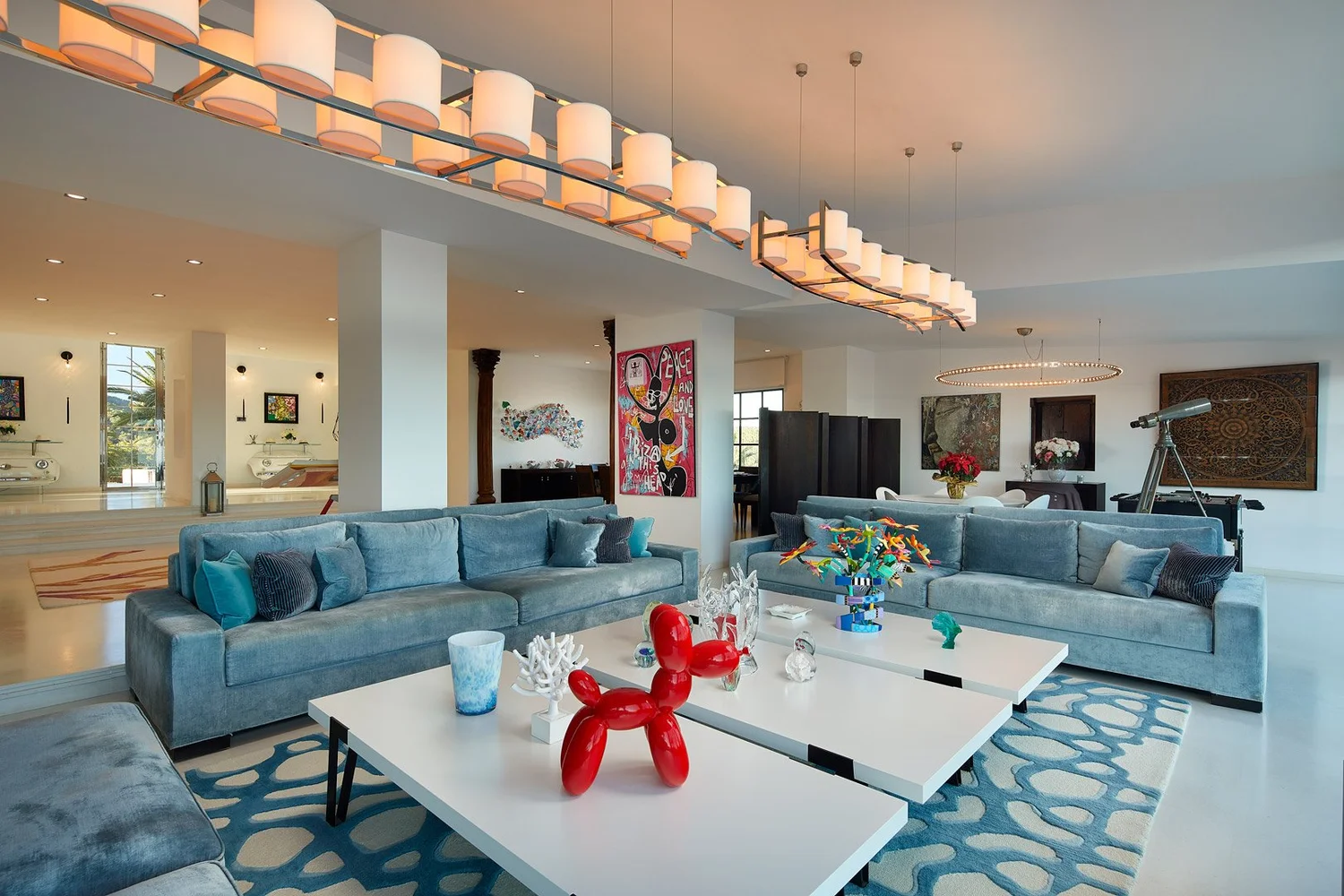 Salón con sofás azules, mesas blancas y obras de arte bajo dos lámparas lineales.