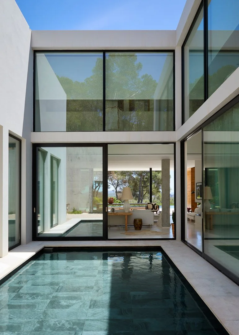 Piscina del patio bajo ventanales de doble altura, con el salón al fondo durante el día.