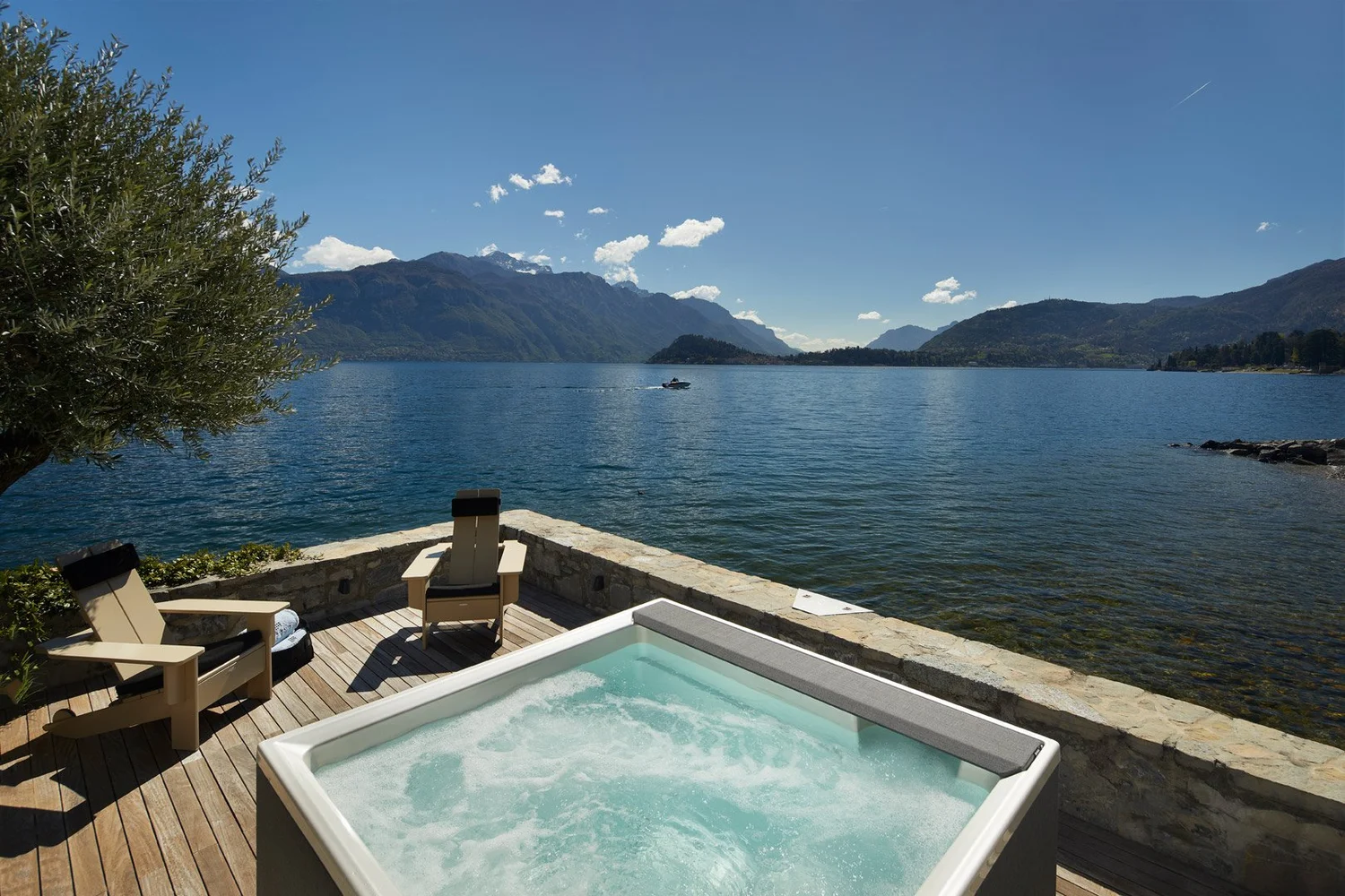 Bañera de hidromasaje y sillones en una terraza junto al lago, con montañas al fondo.