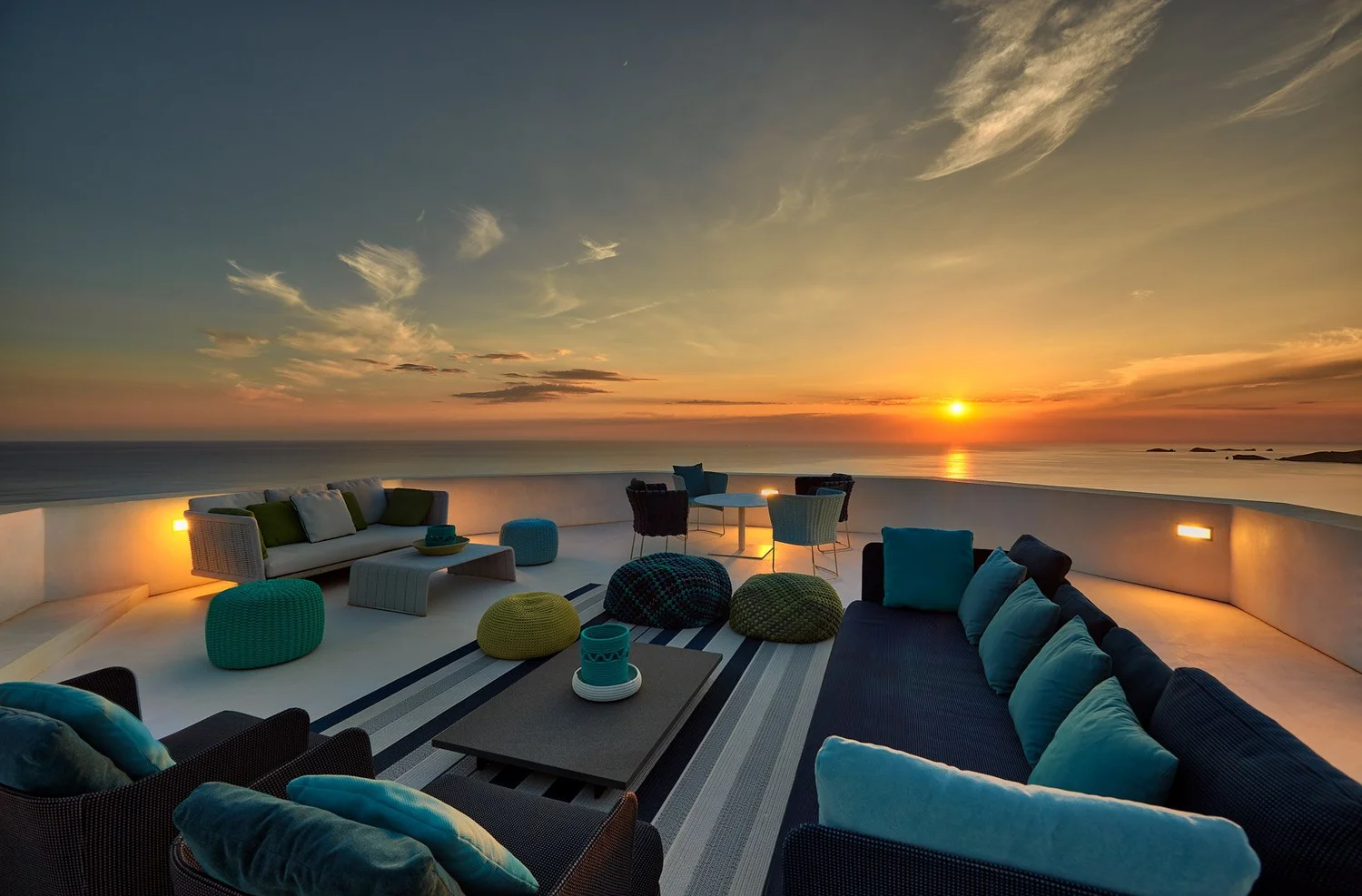 Zona de estar exterior con sofás azules y vistas a la puesta de sol sobre el mar.