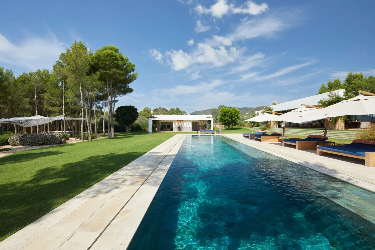 Vista longitudinal de la piscina, con pinos a un lado y tumbonas bajo sombrillas al otro.