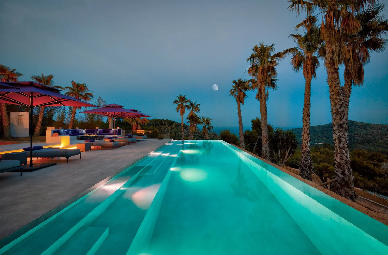 Piscina iluminada al anochecer junto a una hilera de palmeras, con la luna sobre el paisaje.