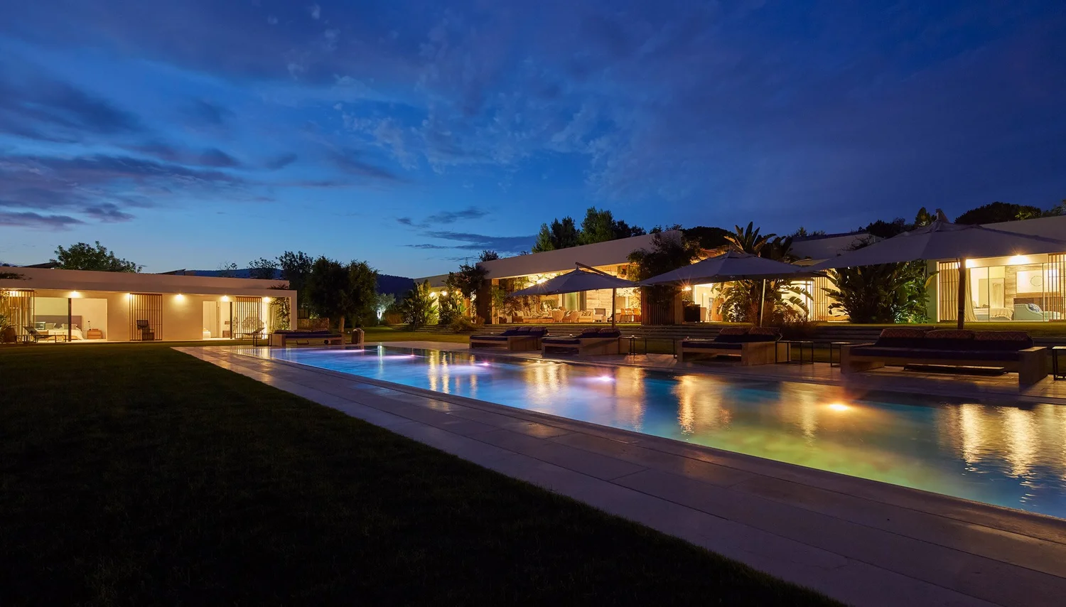 Vista lateral de la piscina y las terrazas iluminadas bajo un cielo azul al anochecer.