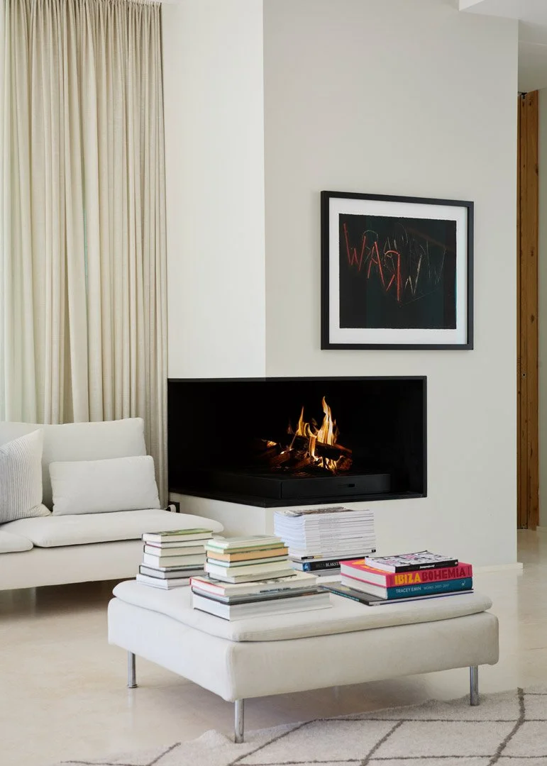Chimenea de esquina encendida junto a un sofá blanco y un puf con libros apilados.