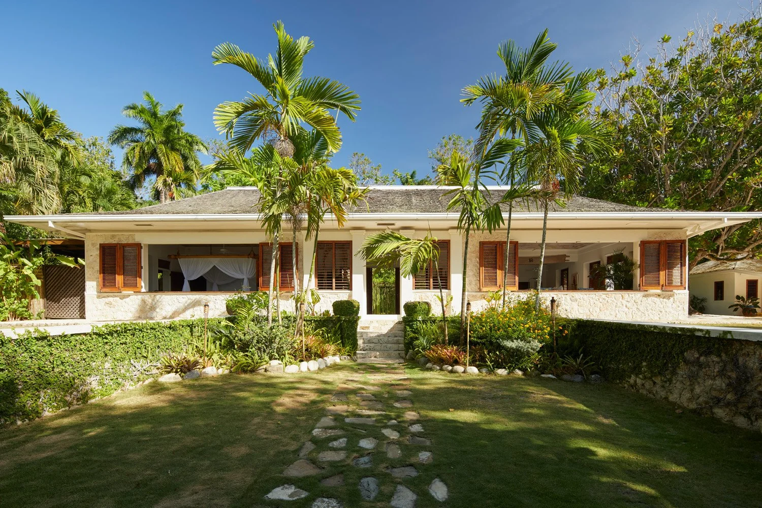 Fachada de la villa durante el día, con contraventanas de madera y palmeras frente al jardín.