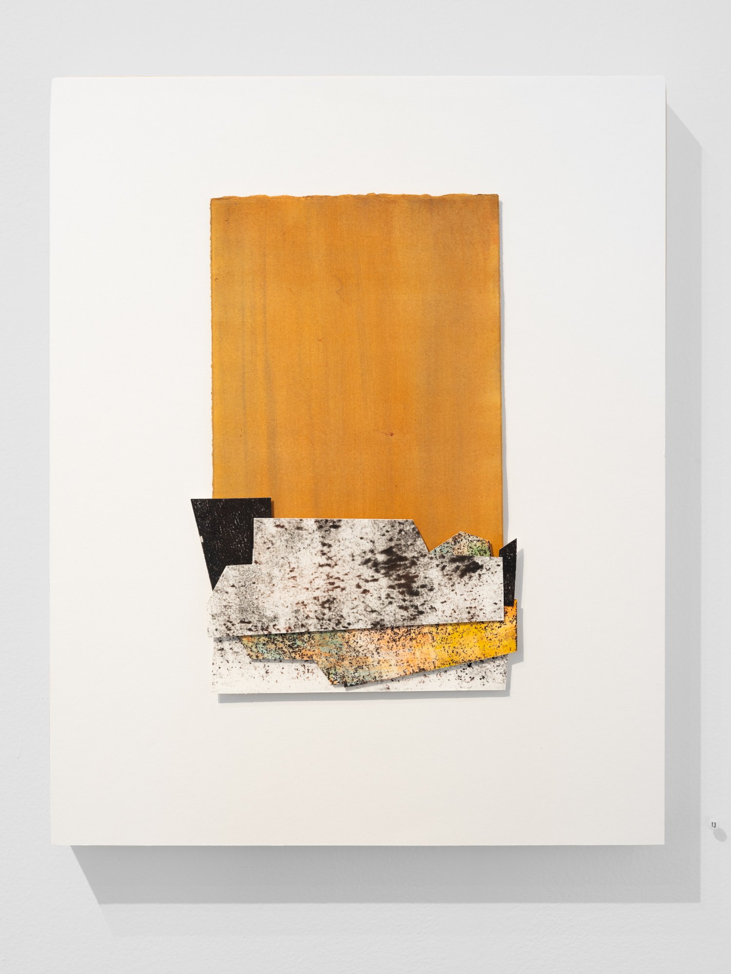Mixed media artwork featuring layered textured paper shapes in earthy tones, mounted on a white background, with a large golden rectangle and various smaller black, white, and speckled paper pieces.