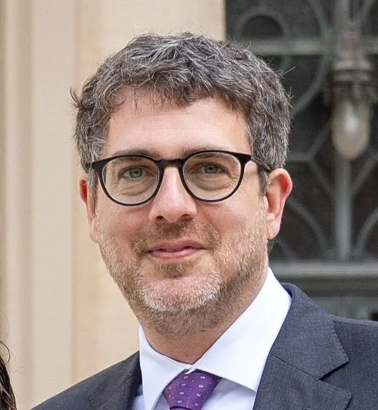 A professional headshot of Avi Cieplinski. The image depicts a middle aged white man wearing a gray suit and purple tie. He has an optimistic expression on his face and wears black glasses. He looks very professional.