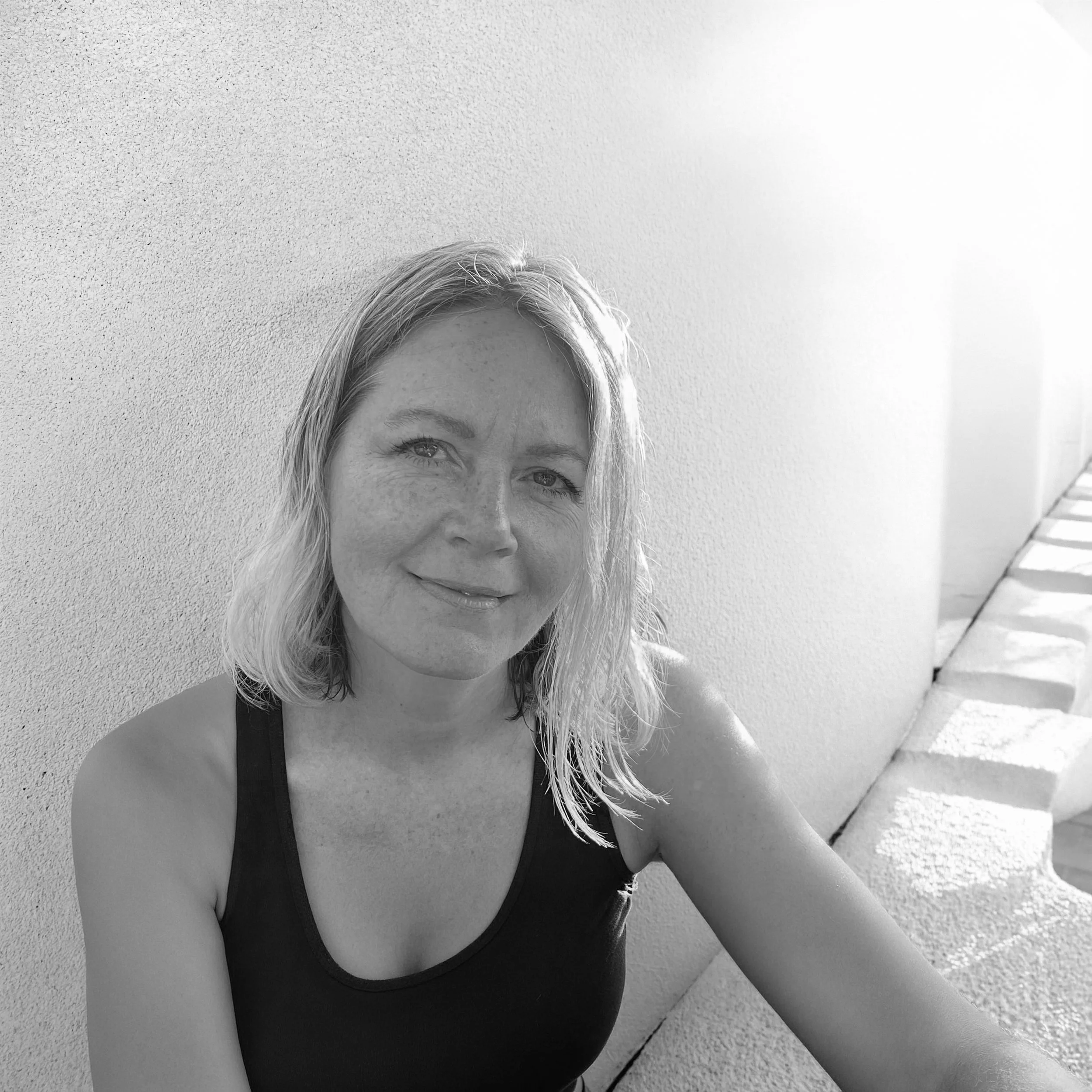 A woman with blonde hair and a smile, wearing a black tank top, sitting outside against a textured wall with steps on the right.