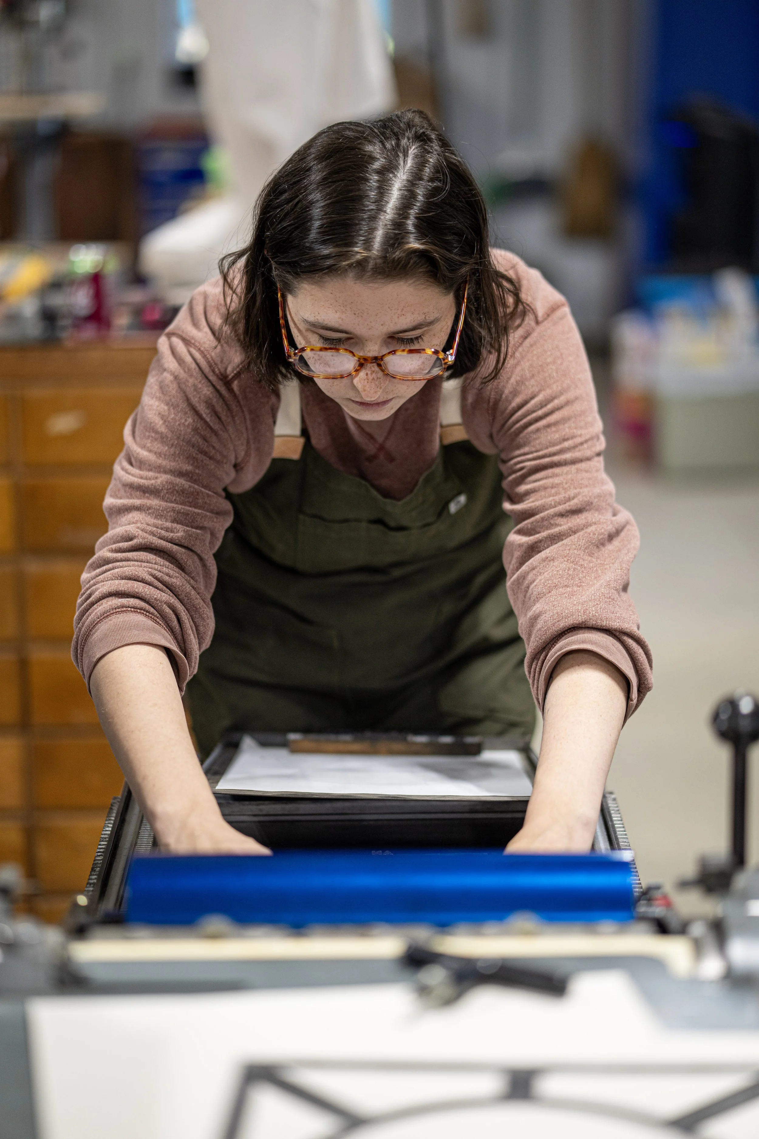 Twenty year old woman with dark hair and glasses looks back at her hands while working on a printing press.