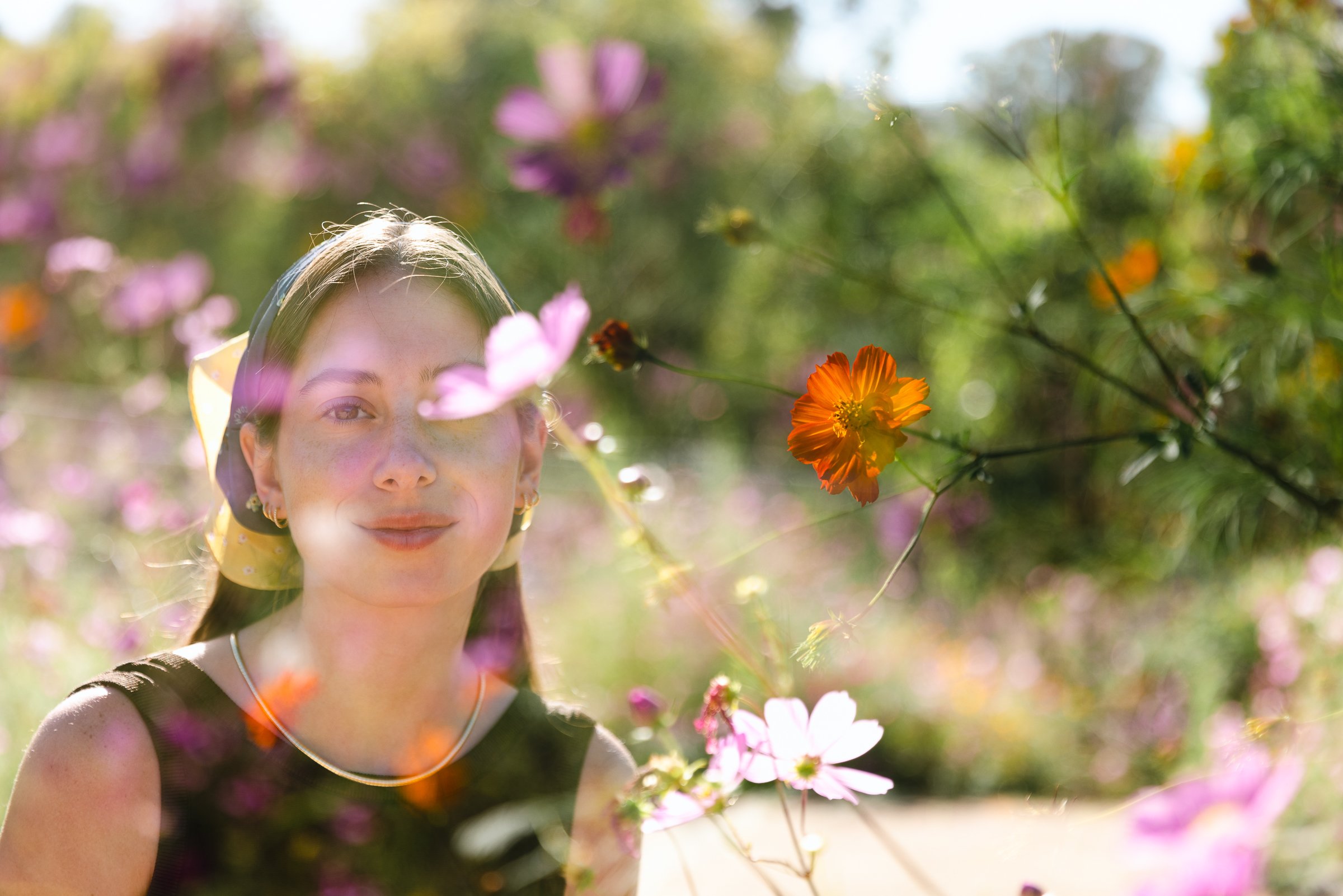 A woman in a garden surrounded by colorful flowers, including pink and orange blossoms, with a blurry natural background.