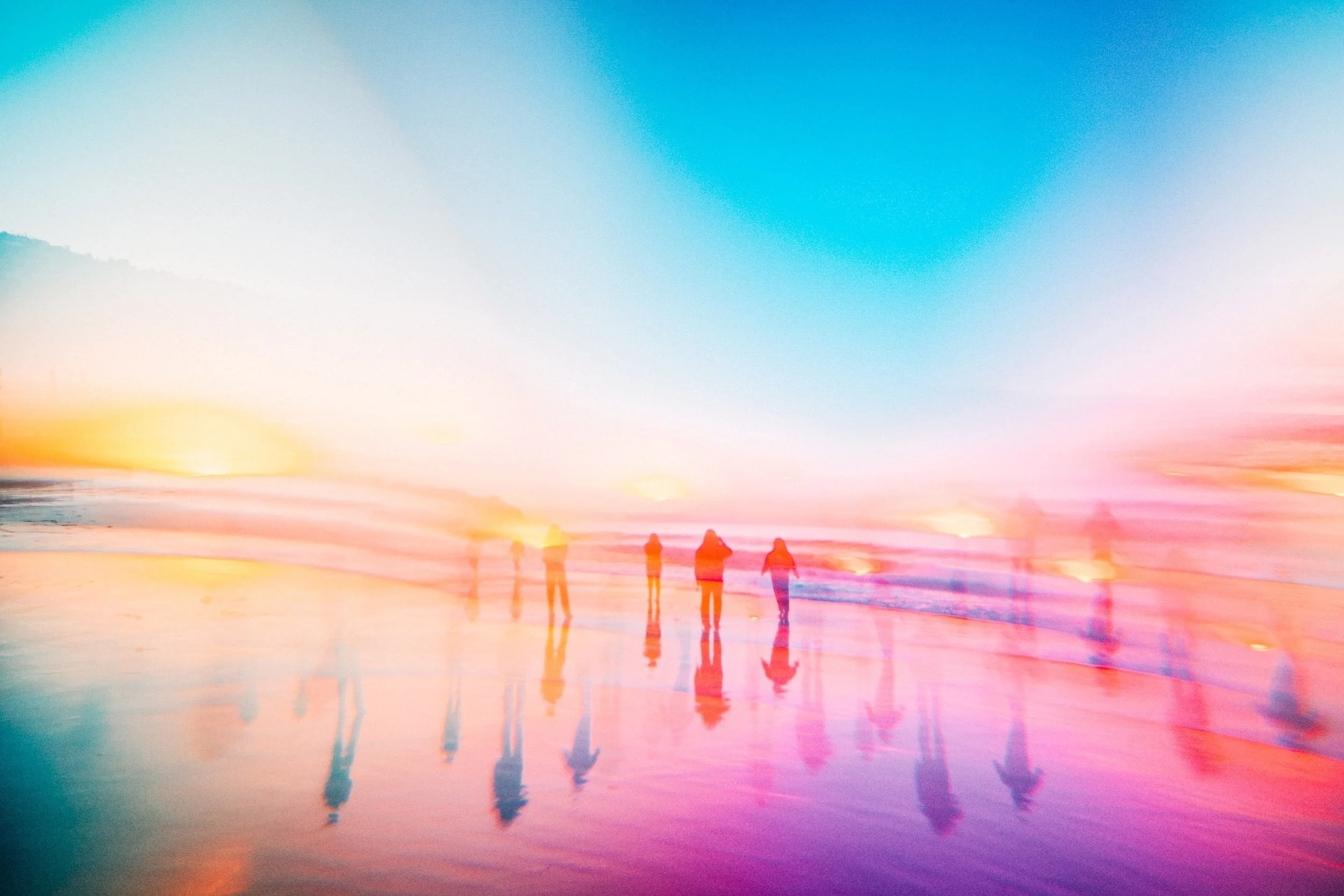 Group of people on the beach in soft rainbow light with a prism-created reflection in the sand.