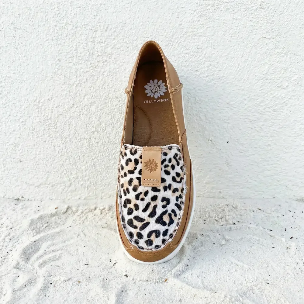 A single tan slip-on shoe with a leopard print fabric on the top part, placed on white sand with a white textured wall background.