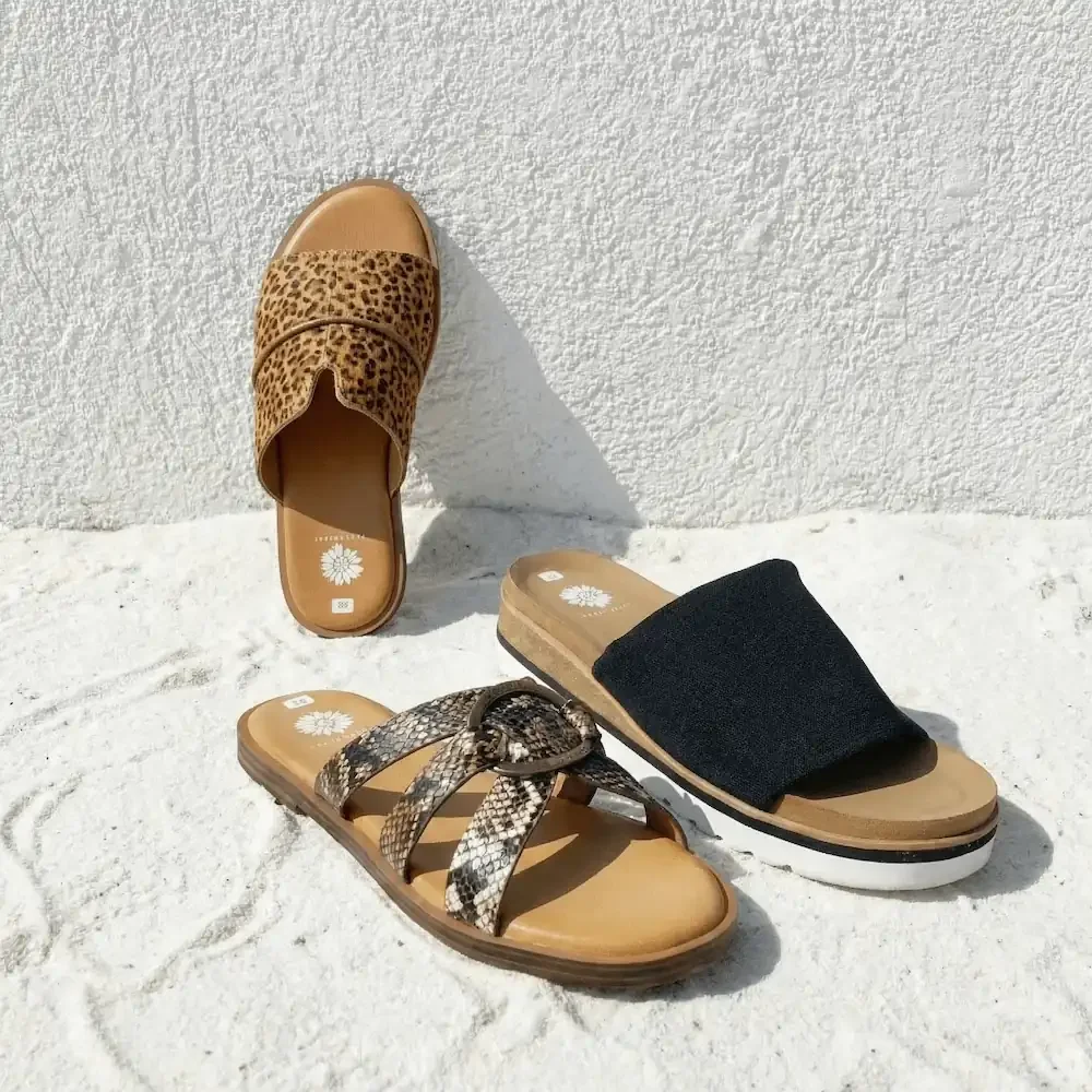 Three pairs of summer sandals on white sand against a white textured wall. One pair has leopard print straps, another has snakeskin straps, and the third has black fabric straps.
