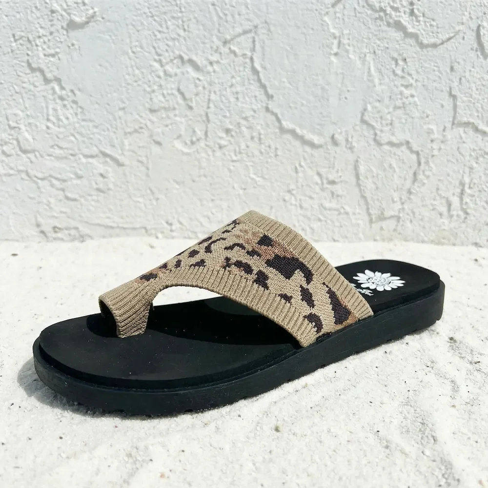 A black flip-flop sandal with a tan strap featuring a leopard print pattern, placed on white sand against a textured white wall.