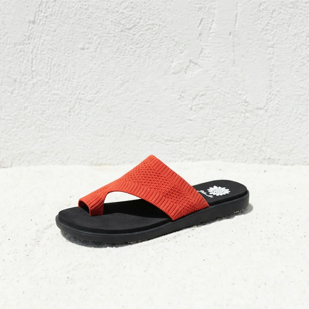 A single flip-flop sandal with a black sole, red strap, and white flower design on the heel, placed on a white textured surface against a white textured wall.