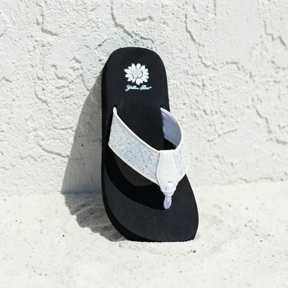 A single black flip-flop with a white glittery strap, leaning against a white textured wall and positioned on sandy ground, with a shadow cast to the left.
