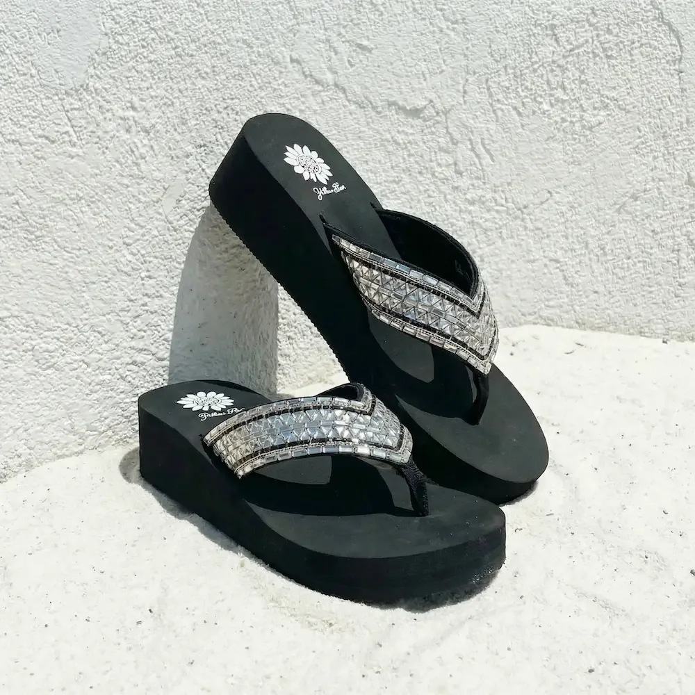 Black flip-flops with rhinestone embellishments on the straps, placed on a white surface against a textured white wall.