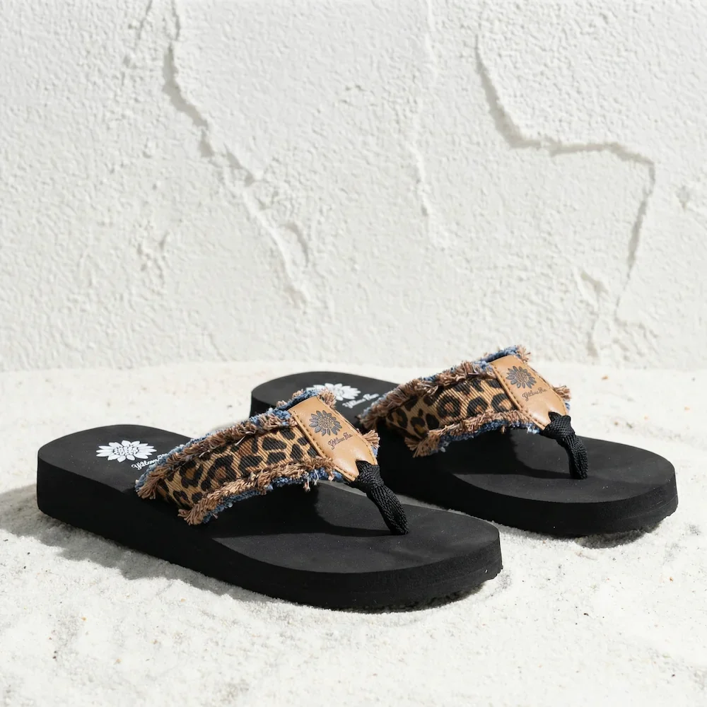 Pair of black flip-flops with frayed leopard print straps on a sandy surface.