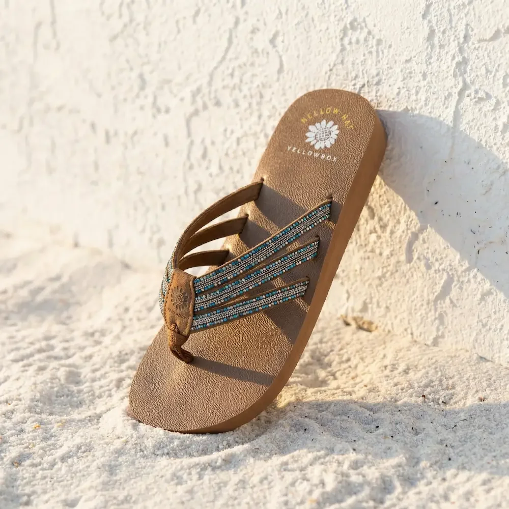 Beige flip-flop sandal with blue and white beaded straps, leaning against a white textured wall on sand.