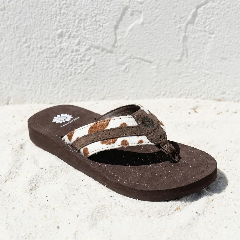 A brown flip flop sandal with a white and brown animal print strap resting on white sand against a white textured wall.