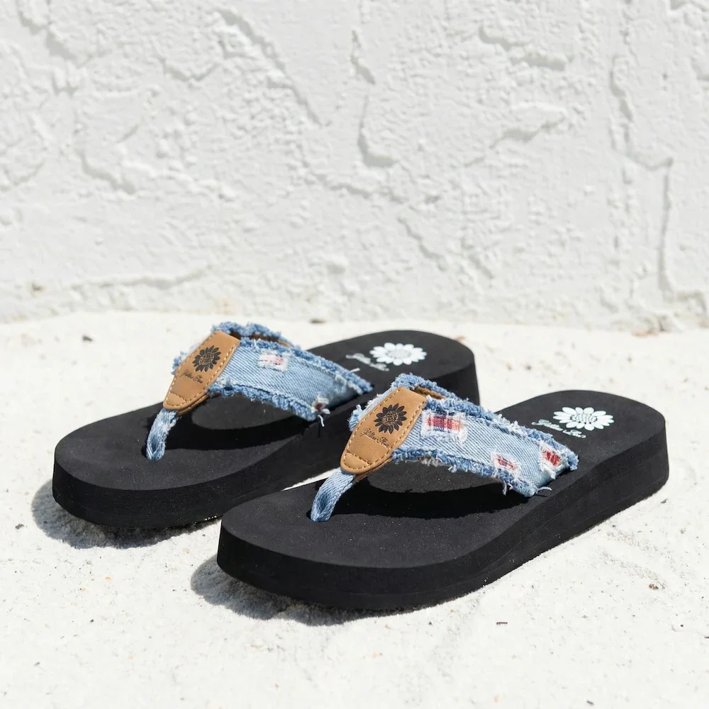 Pair of black flip-flops with denim straps and a small leather tag, placed on white sand with a textured white wall in the background.