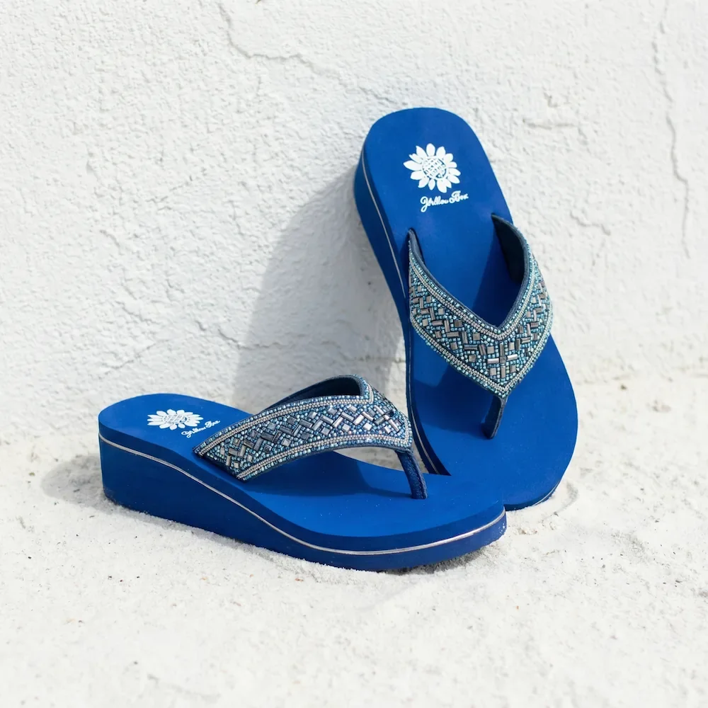 Blue beach flip-flops with rhinestone embellishments on the straps, positioned on white sand against a white textured background.