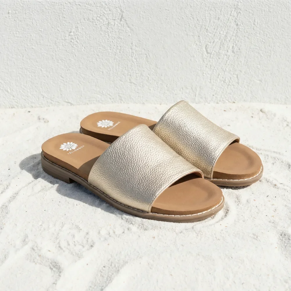 A pair of beige or light gold slide sandals on a white textured surface, with a plain white background.