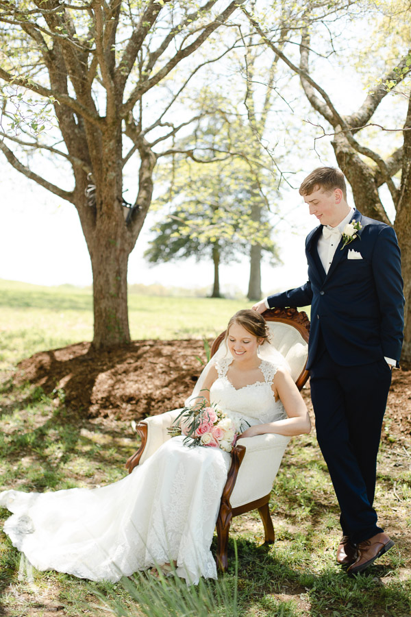 Bride sitting in chair with groom standing by her side.