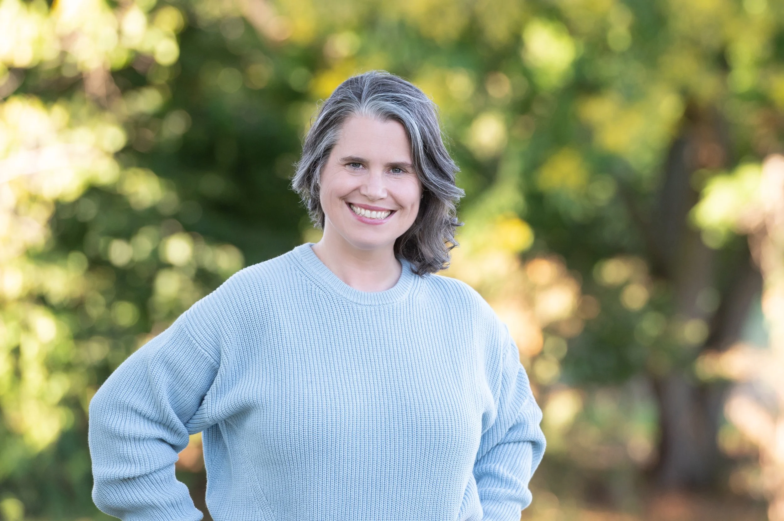 Smiling woman in blue sweater against a tree background. Image was taken at the golden hour.