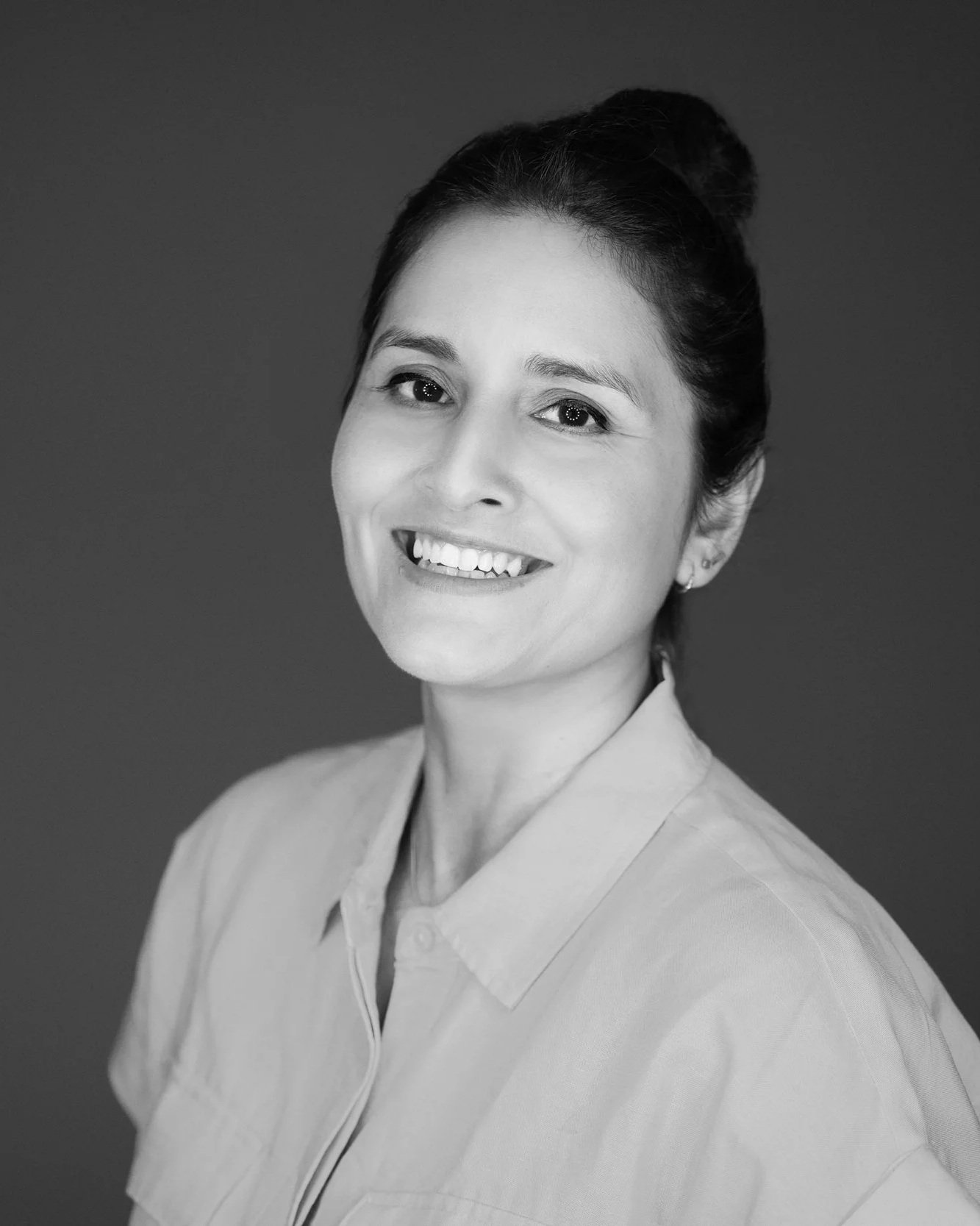 Black and white portrait of a woman with dark hair styled in a bun, smiling, wearing a collared shirt, and earrings.